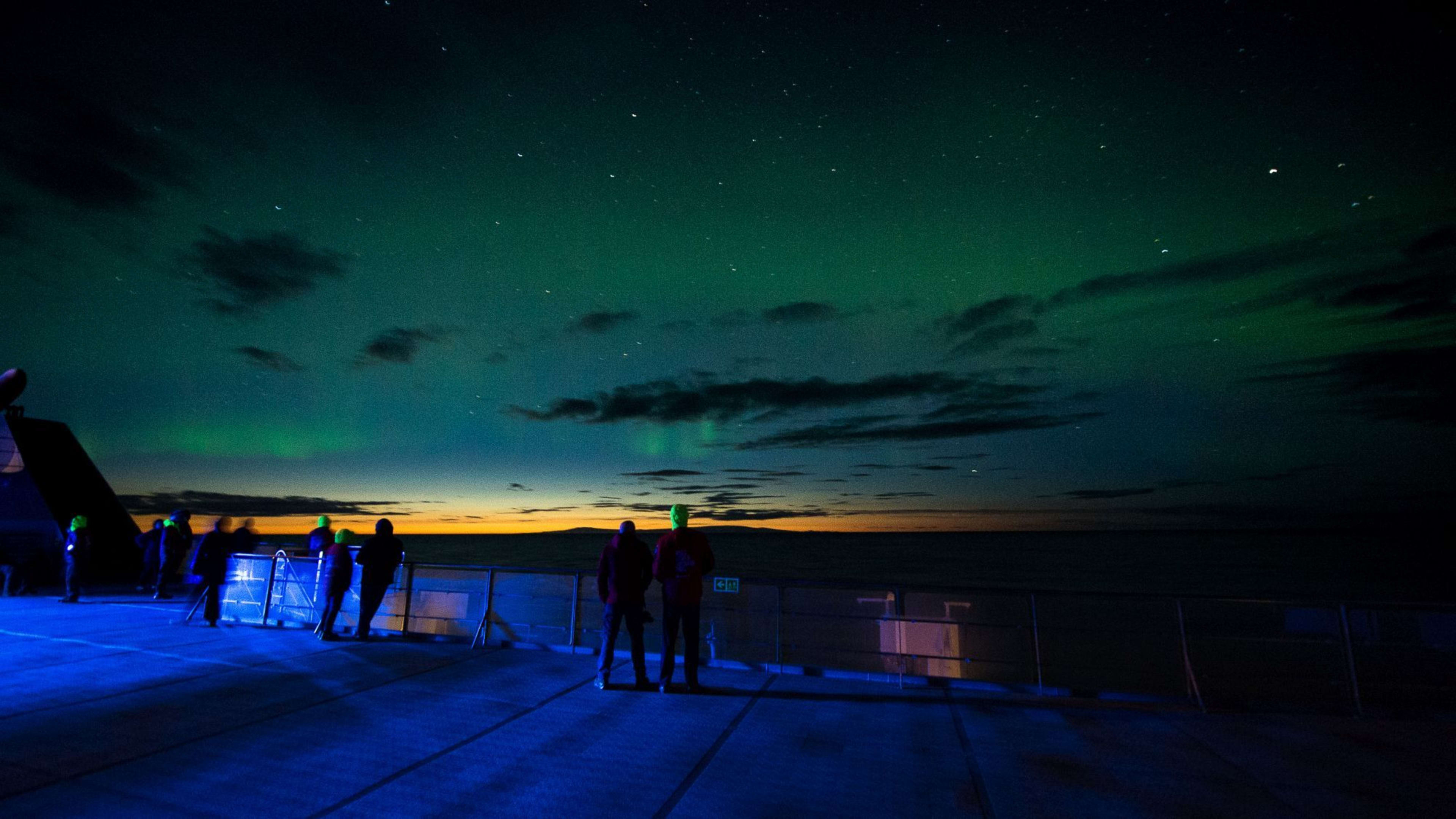Group on ship deck under starry sky with faint Northern Lights above, glowing horizon at dusk, blue lighting on deck.