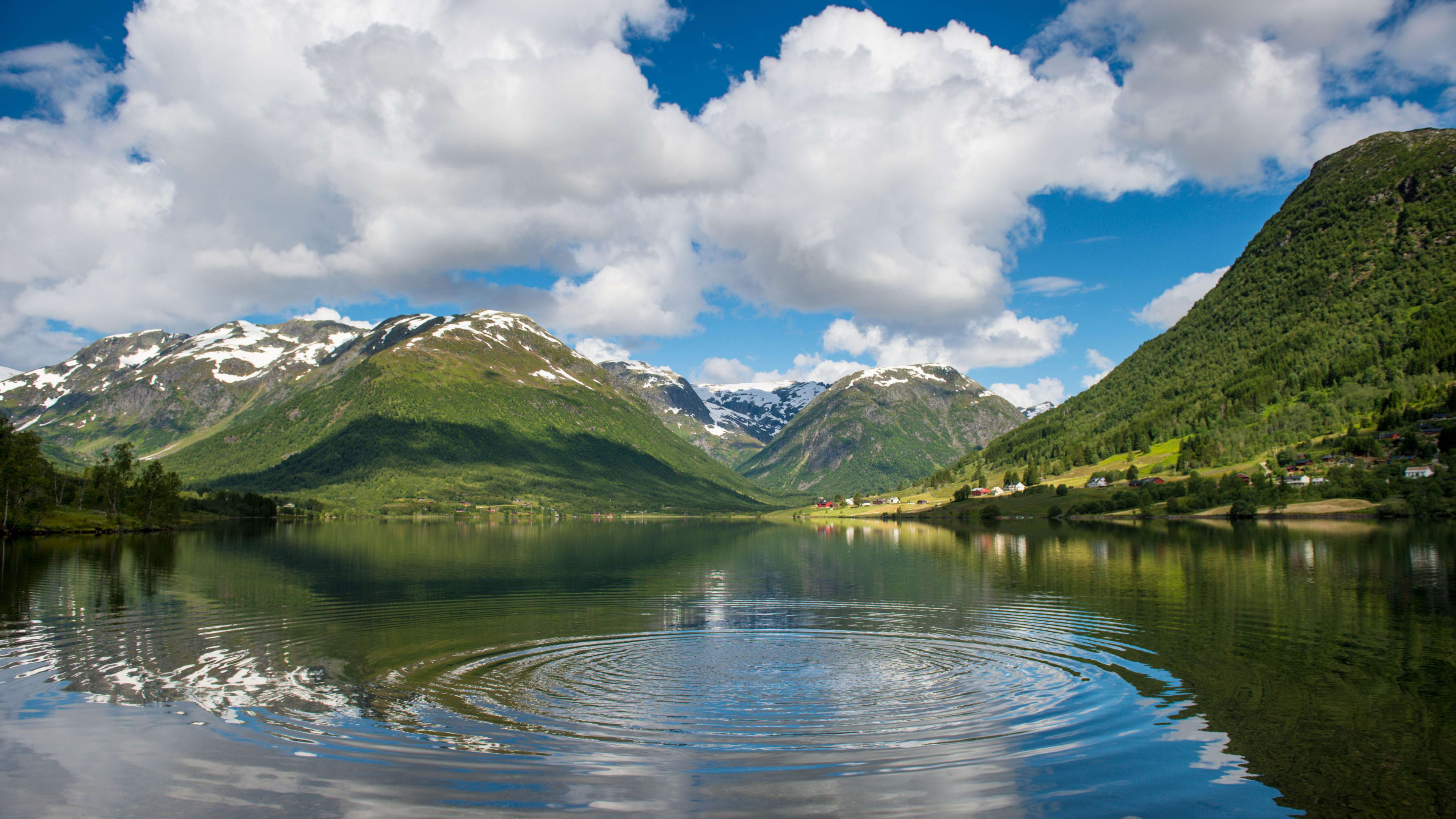 Lake reflects snowy peaks and green hills under sunny sky.