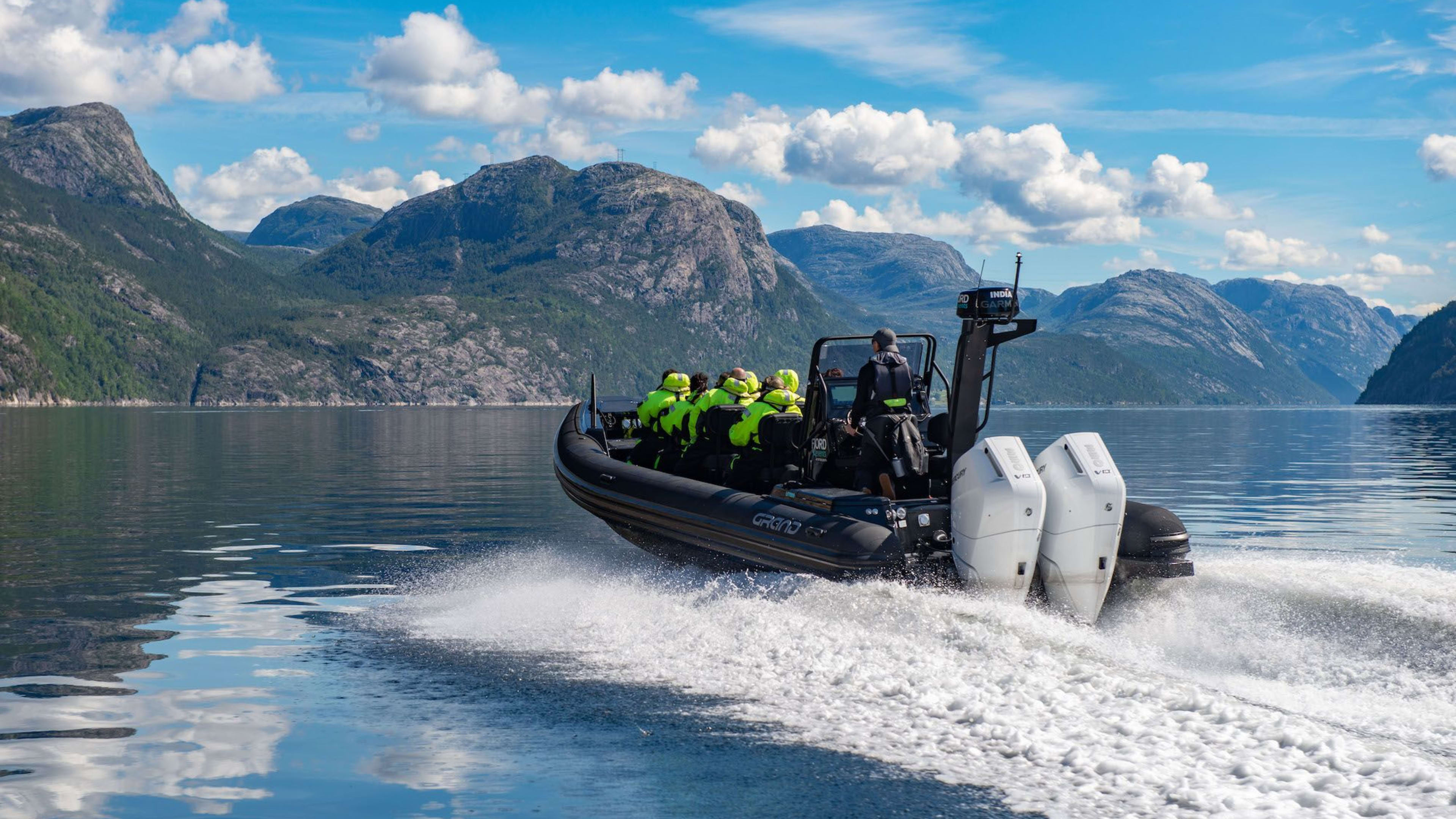 Ein schwarzes aufblasbares Schnellboot fährt über einen ruhigen Fjord, Passagiere in neon-grünen Jacken.