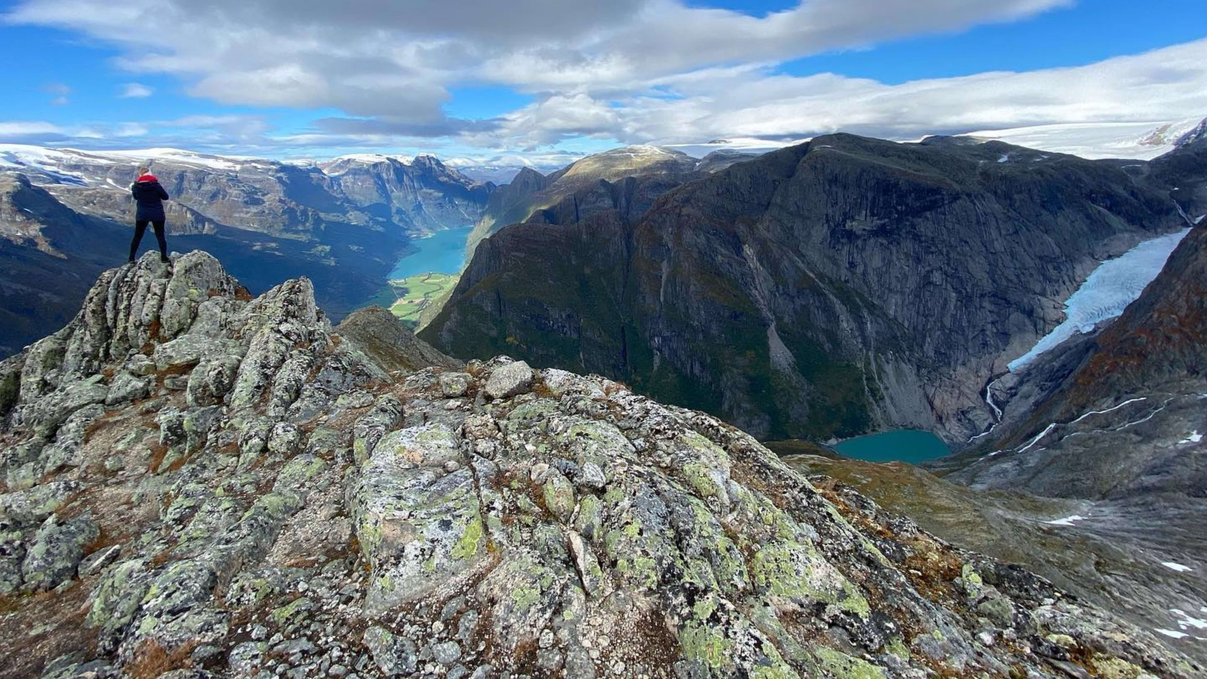 Person som står triumferende på en steinete topp, med utsikt over fjorden og snøkledde fjell i bakgrunnen.