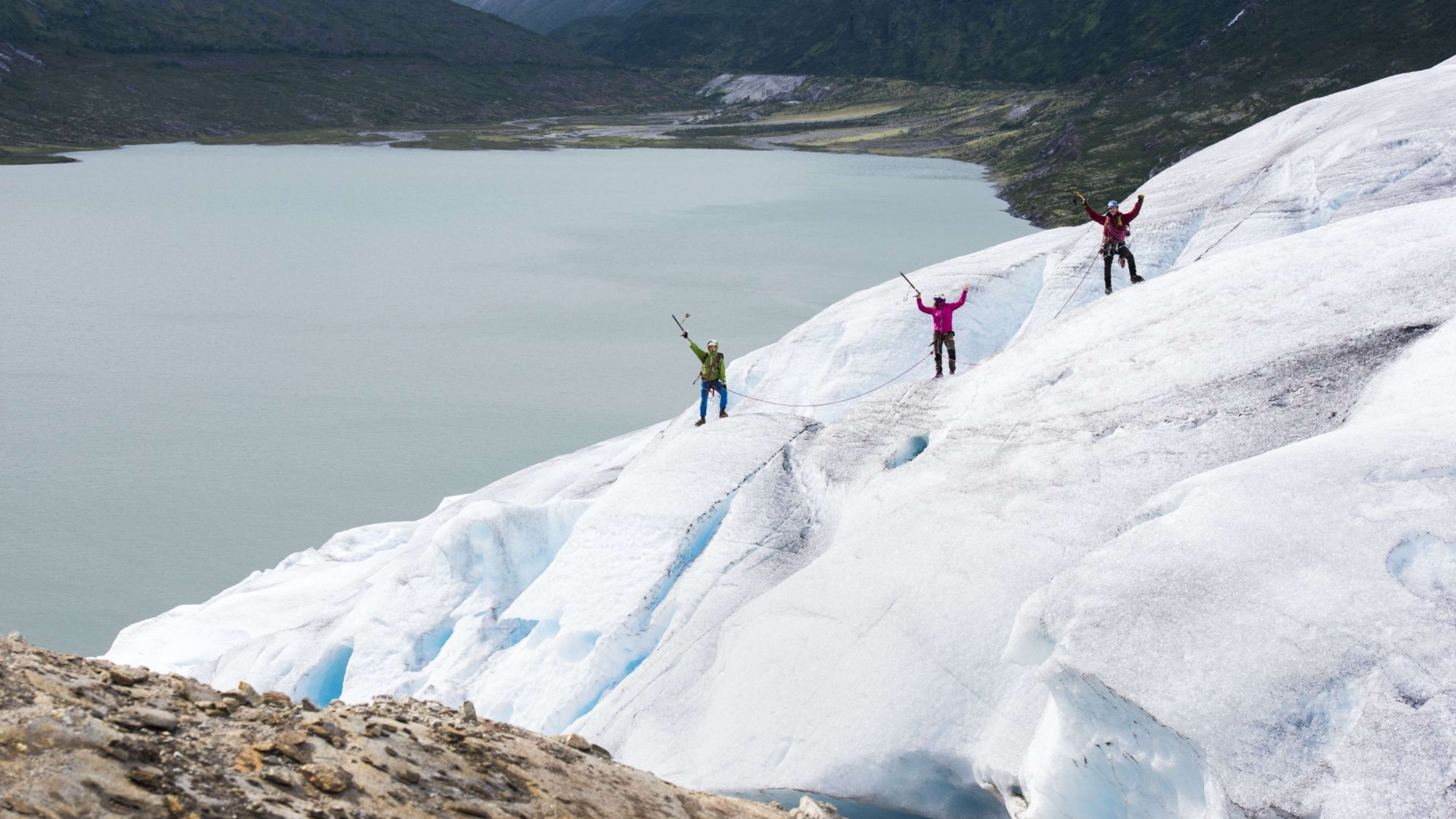 Three climbers on glacier, connected by rope, celebrating on icy surface with fjord and mountains in the background.