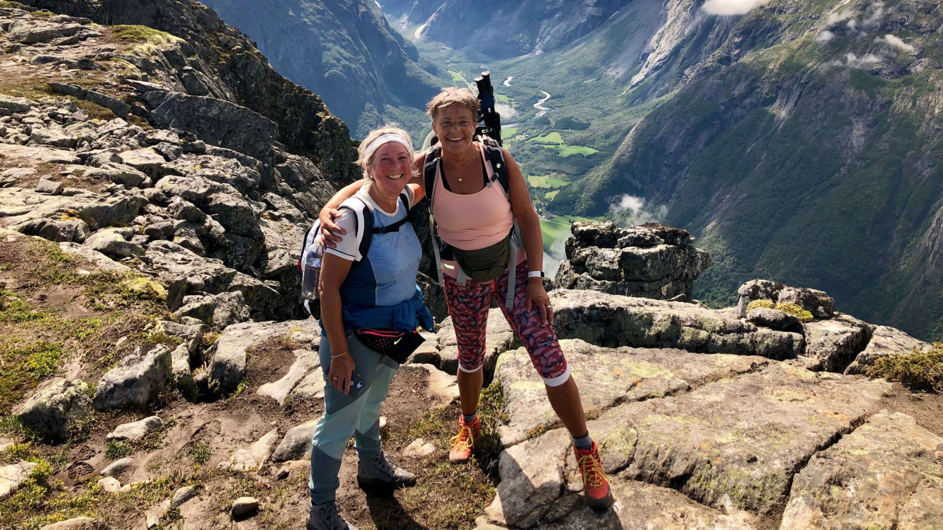 Zwei Frauen umarmen sich auf einem Berg, mit Tal und schneebedeckten Gipfeln in der Ferne.