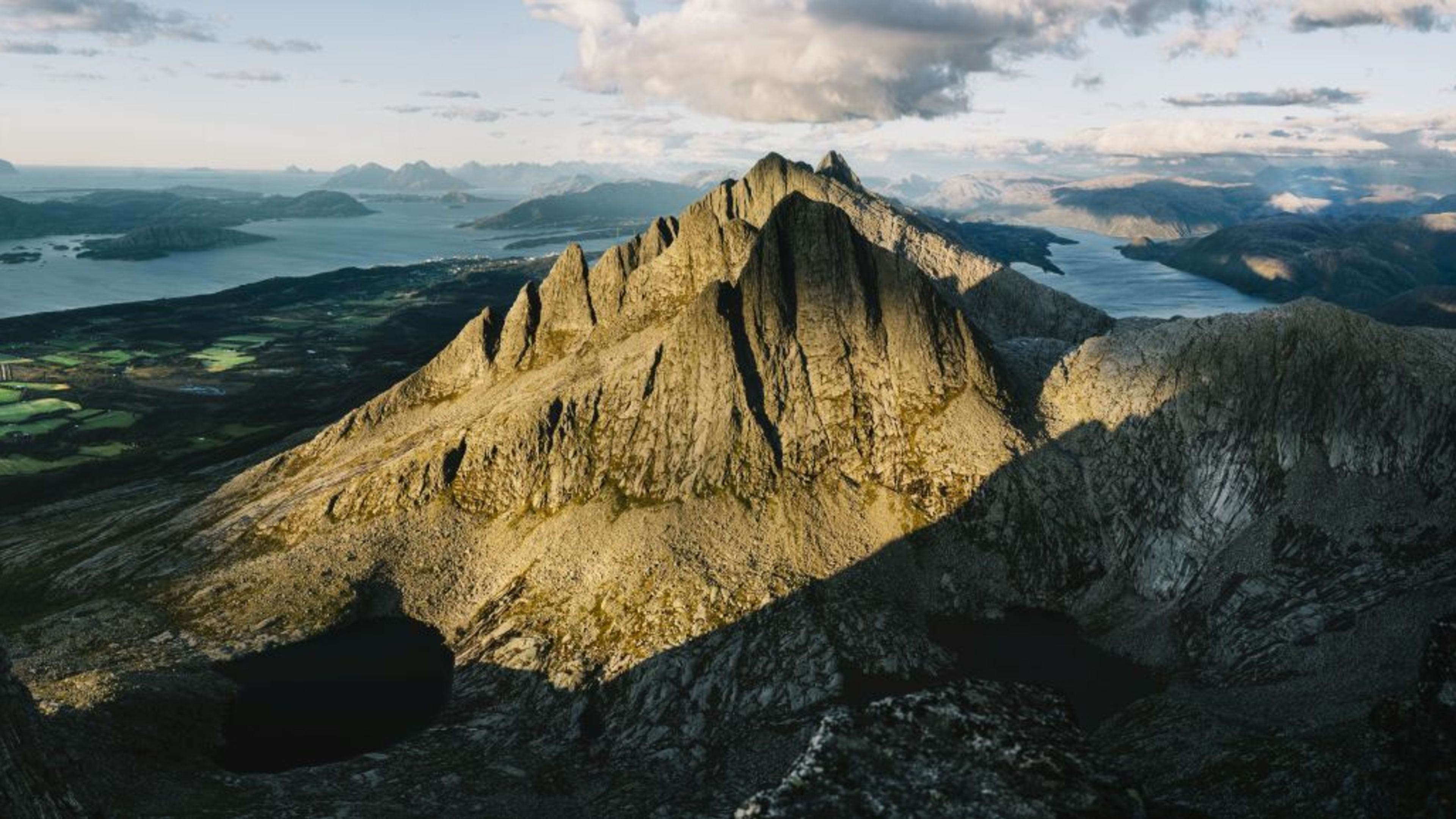 Fotograf in gelber Jacke, der die Küstenlandschaft mit dramatischen Gipfeln, grünen Ackerflächen und ruhigem Meer einfängt.