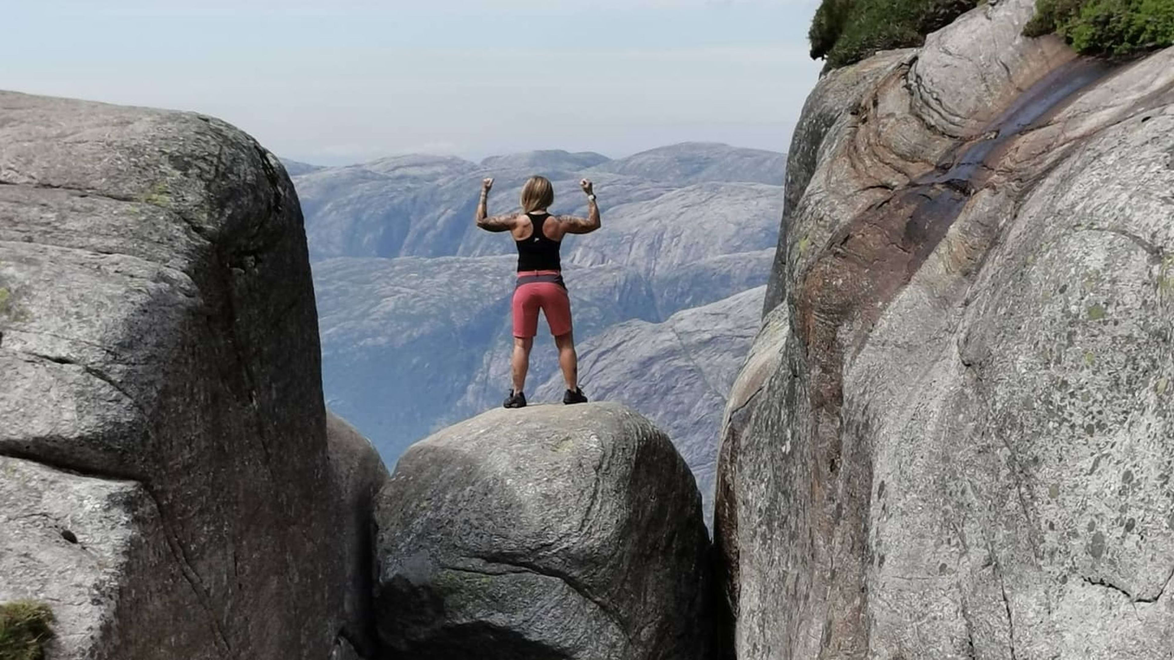 Kvinne som står triumferende på Kjeragbolten, en stor stein som sitter mellom to bratte fjellvegger over en dyp fjelldal.
