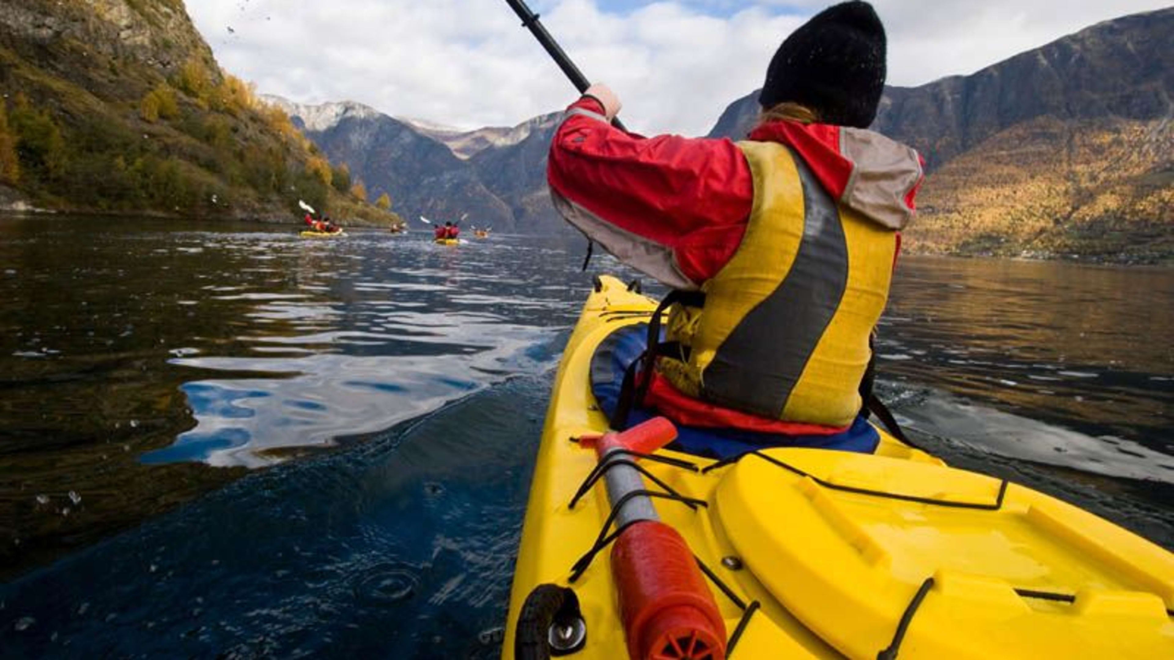 Kajakfahrer, der auf einem ruhigen, dunkelblauen Wasser in einem gelben Kajak paddelt, umgeben von herbstfarbenen Bergen, mit fernen Kajakfahrern im Hintergrund.