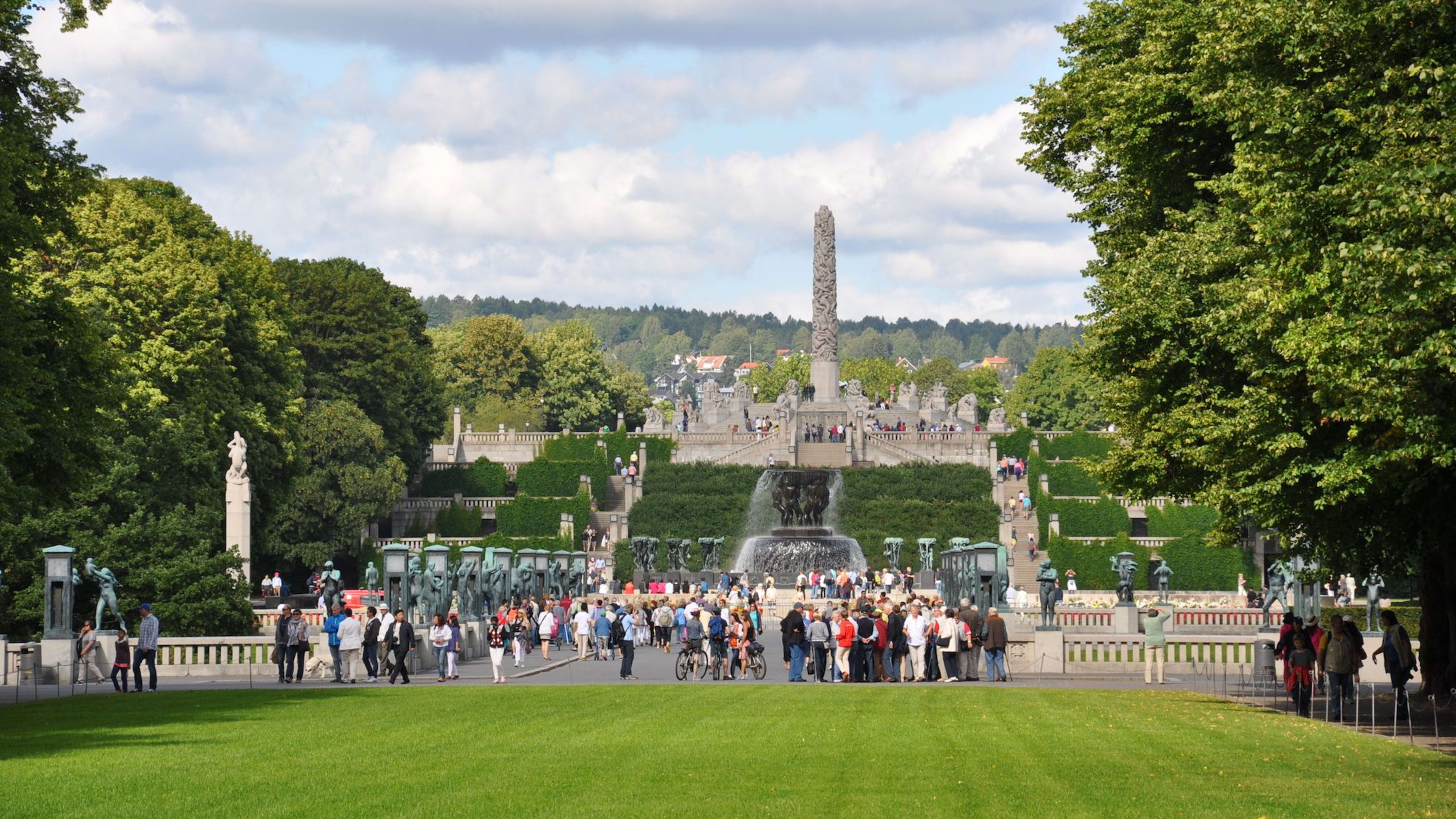 Folkemengder går i Vigelandsparken i Oslo med Monolitten og skulpturer blant grønne trær.