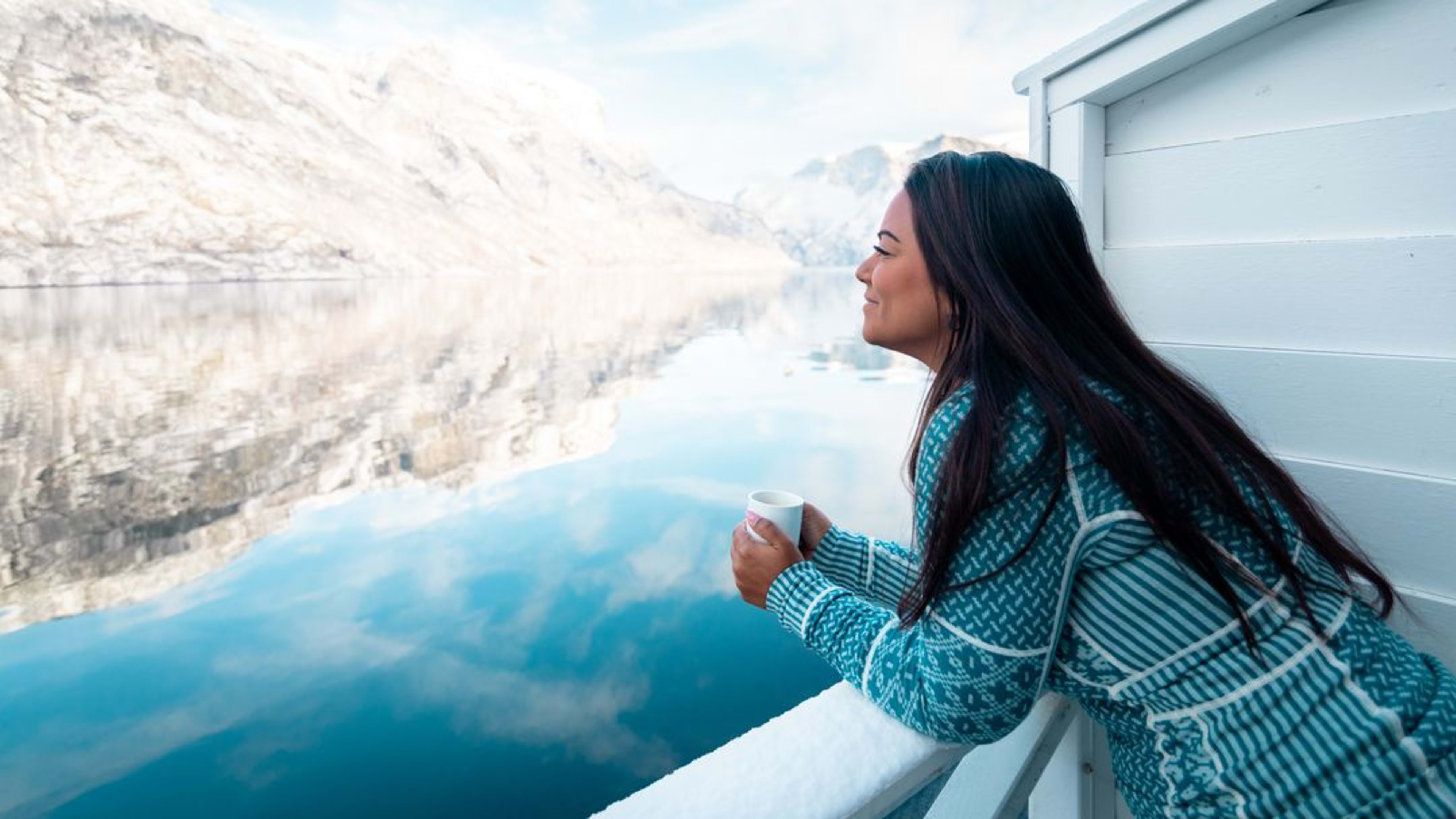 Woman in sweater holds cup, gazes at snowy Nærøyfjord surrounded by mountains.