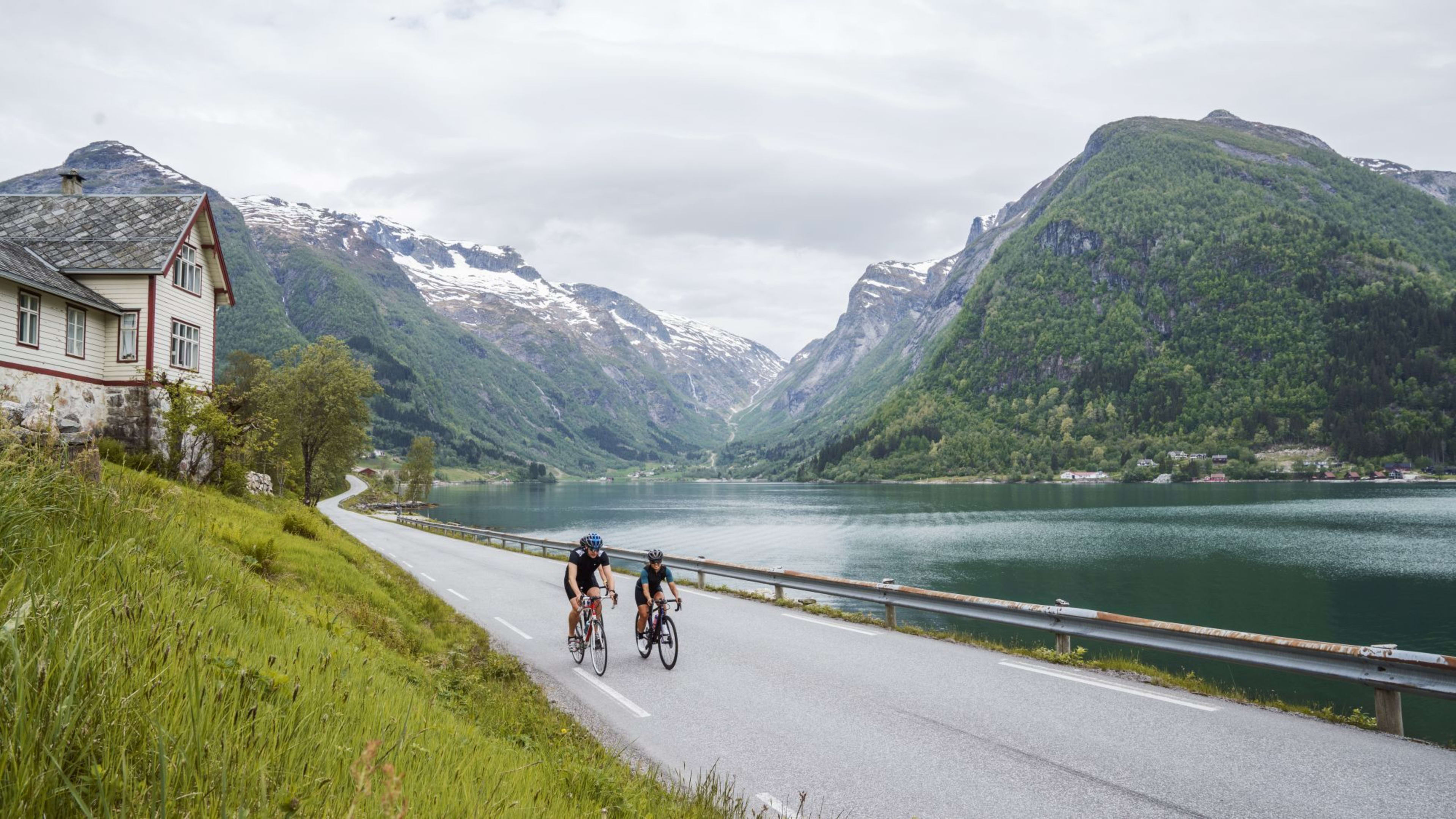 Radfahren in Balestrand am Sognefjord, vorbei an Häusern mit schneebedeckten Bergen im Hintergrund.