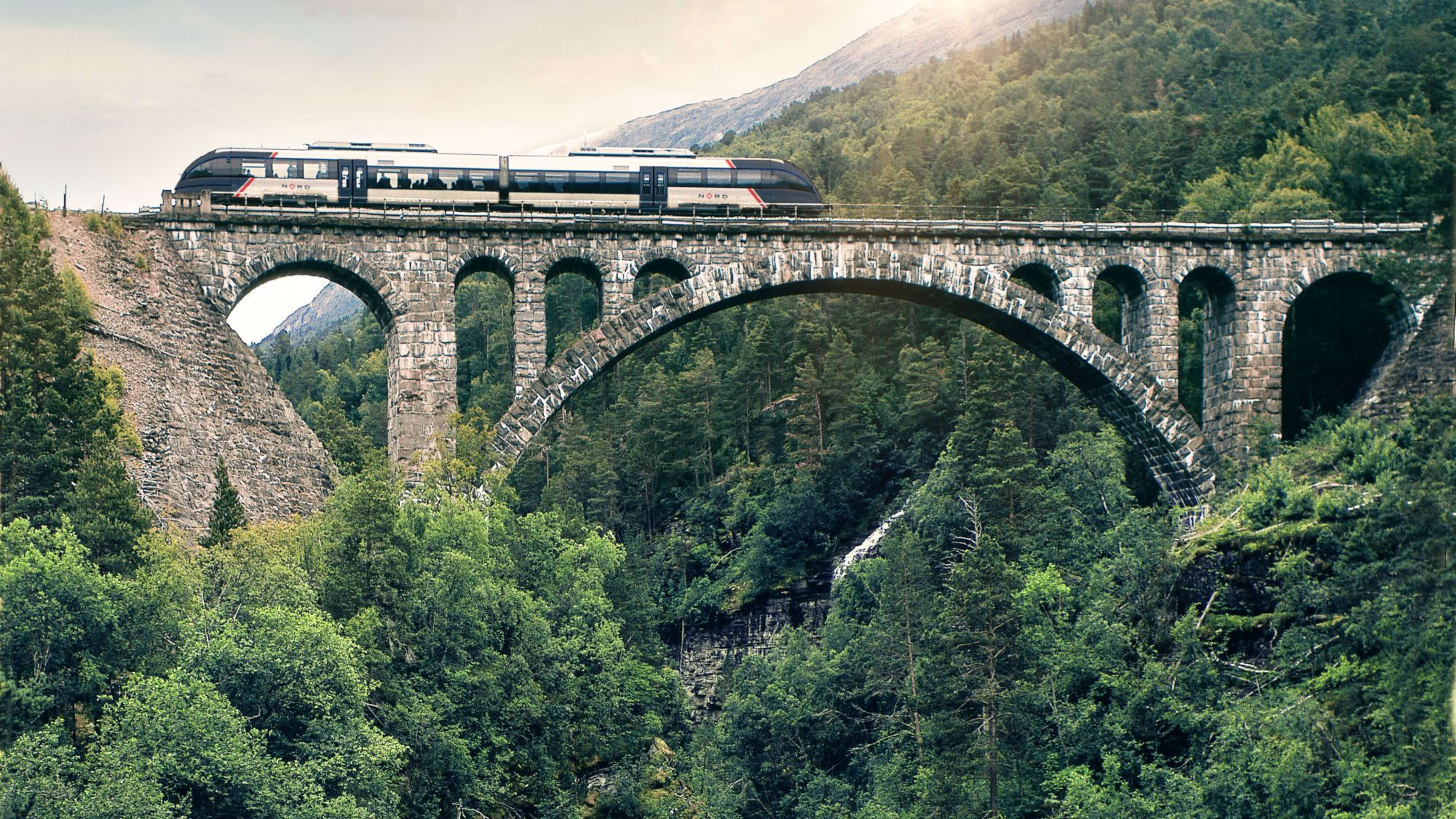 Train crosses old stone bridge over gorge with river, sun breaks through cloudy sky.