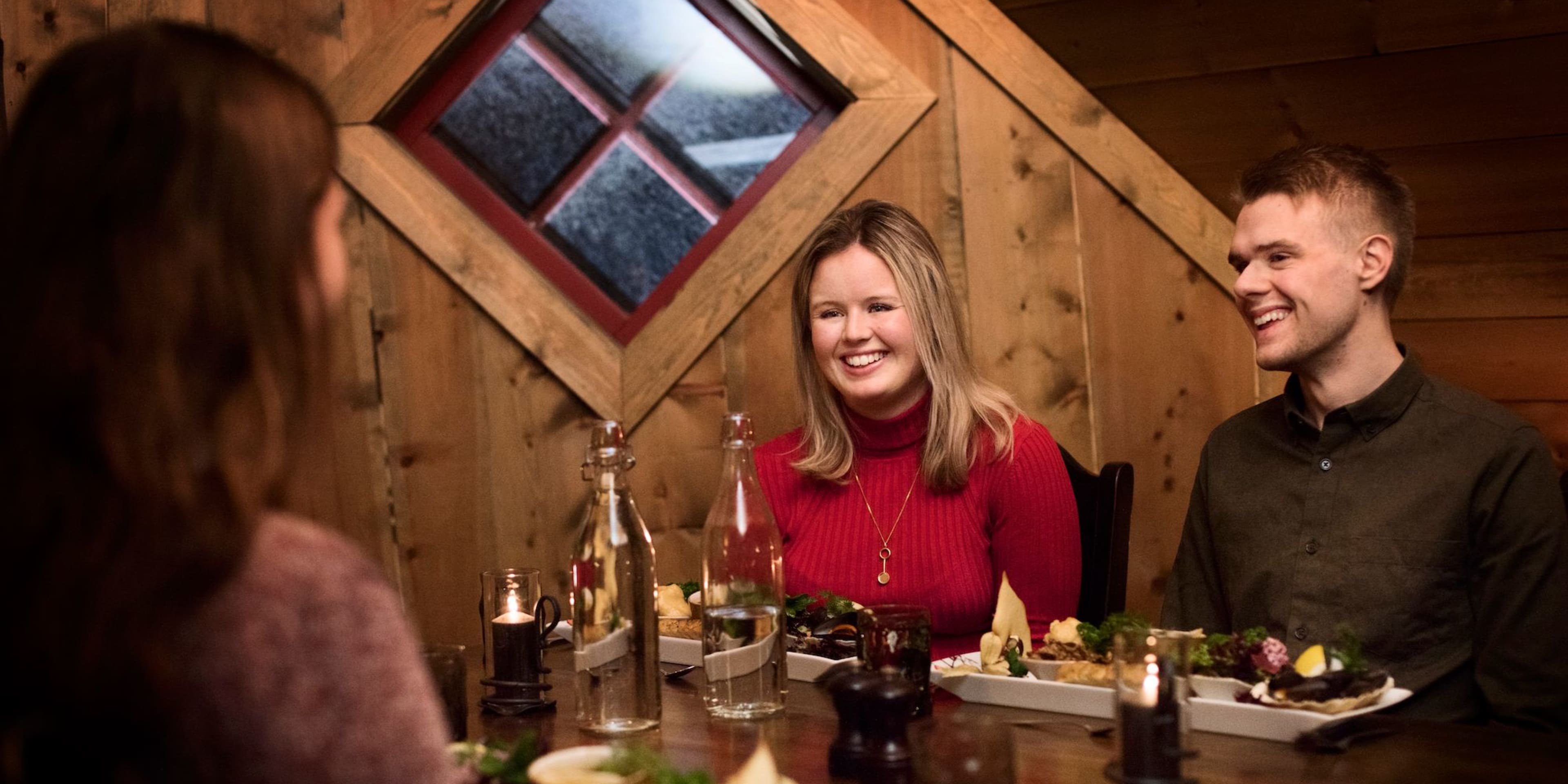 Freunde genießen ein gemütliches Abendessen in einer rustikalen Holzhütte mit Tellern mit Essen, Kerzen und einladender Atmosphäre.