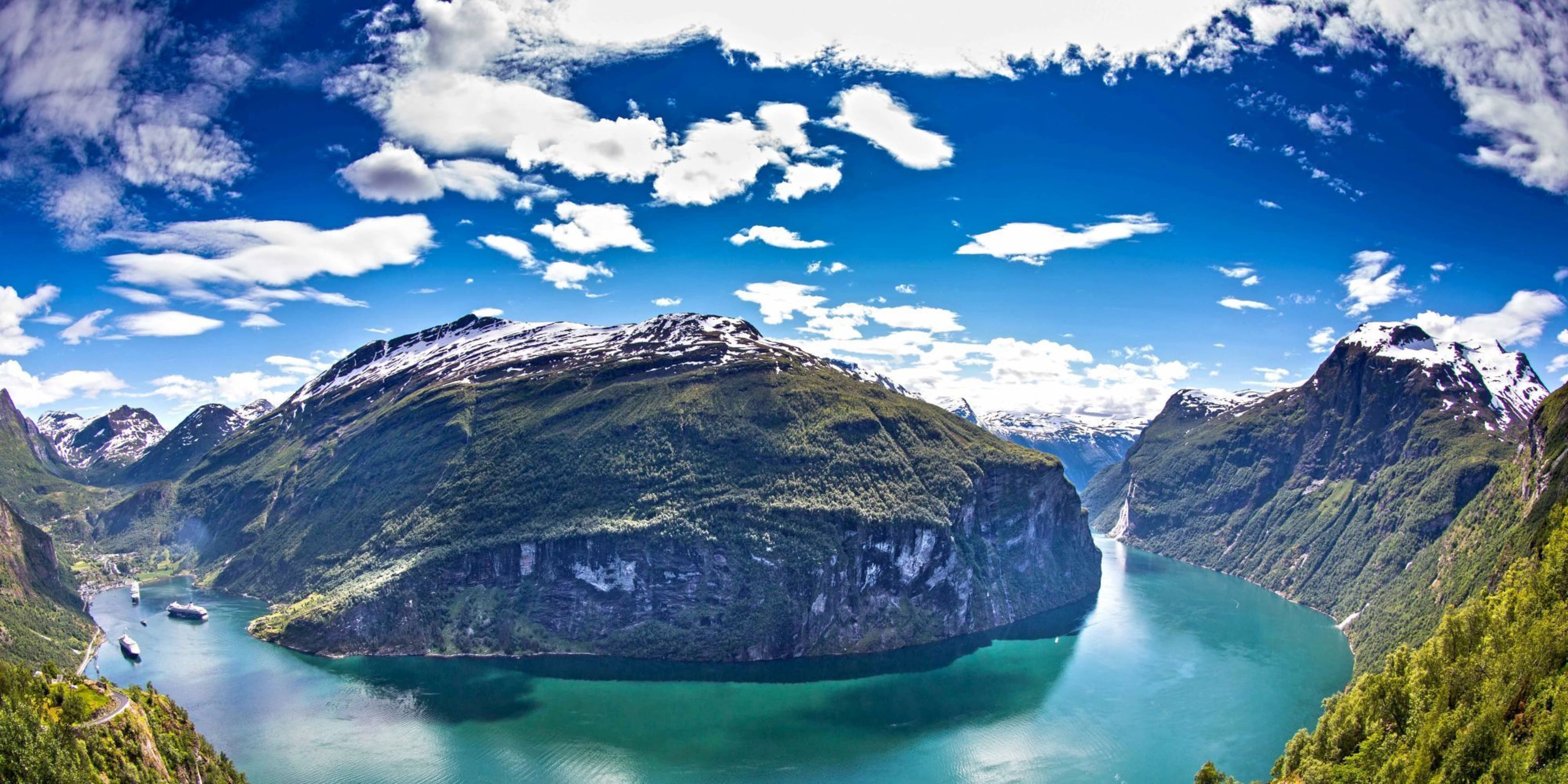 Turquoise fjord with cruise ships, forested mountains and snow.