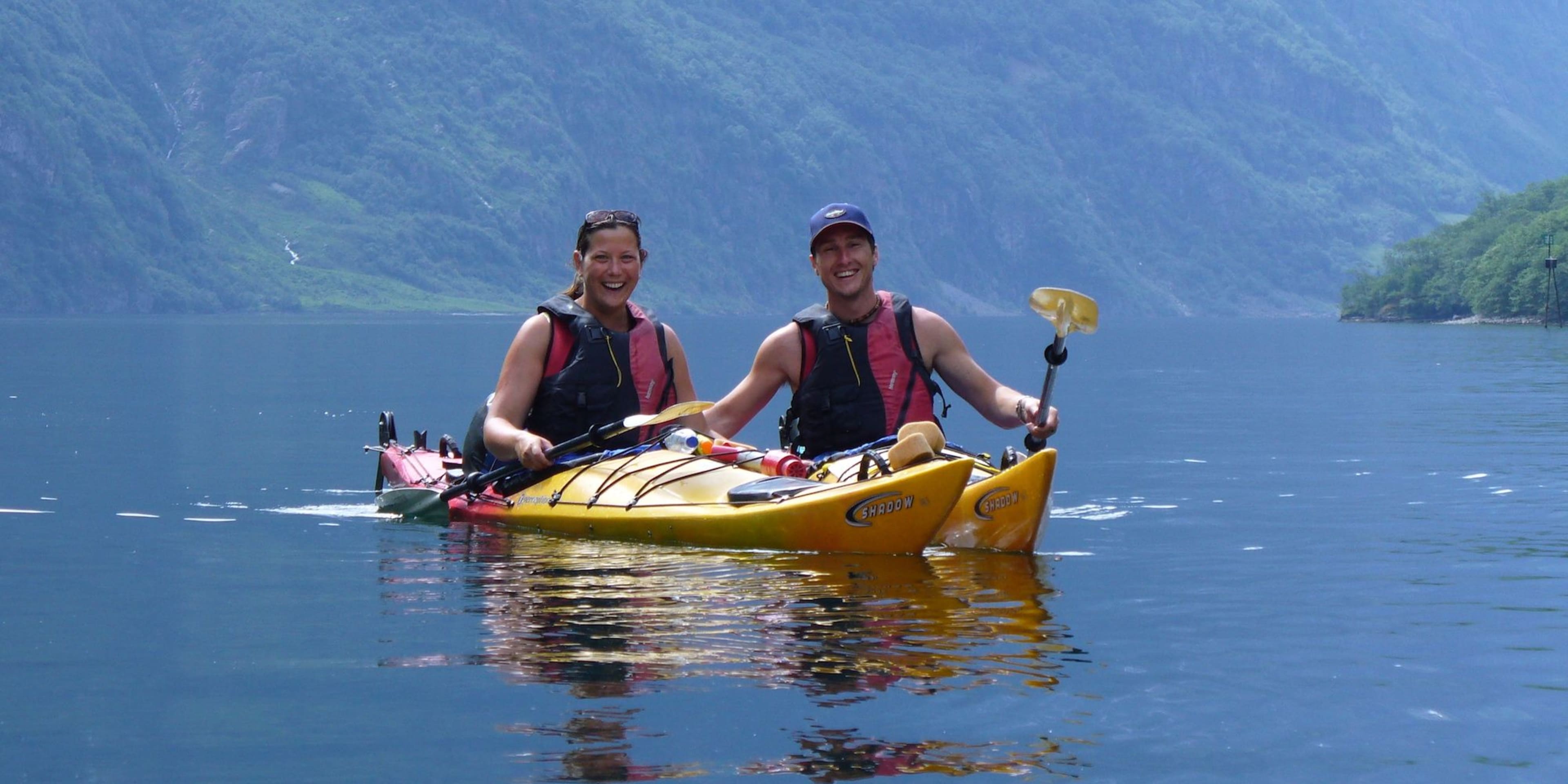 Two smiling kayakers in a yellow tandem kayak on calm blue water, surrounded by green hills and waterfalls in a peaceful fjord.