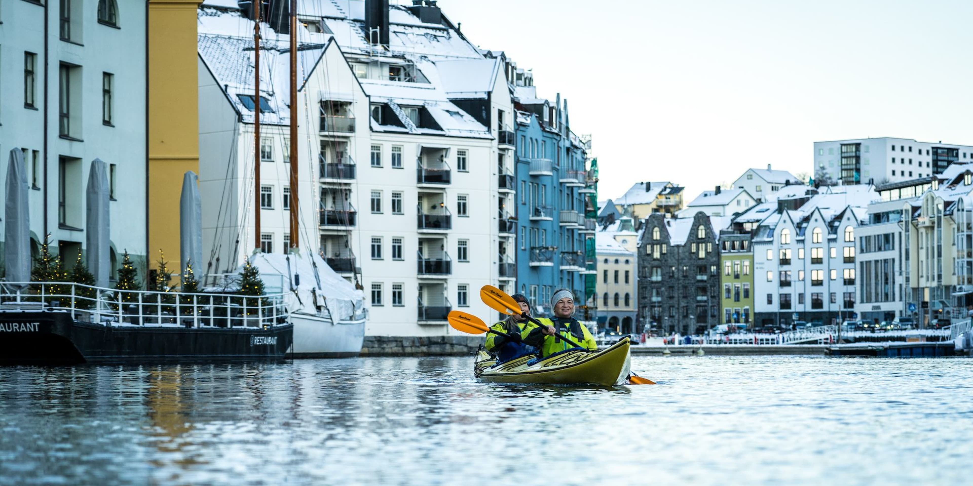 Zwei Personen paddeln in der Nähe von bunten, schneebedeckten Gebäuden, mit einem Boot, das in der Nähe angedockt ist, und festlichen Dekorationen entlang des Docks.