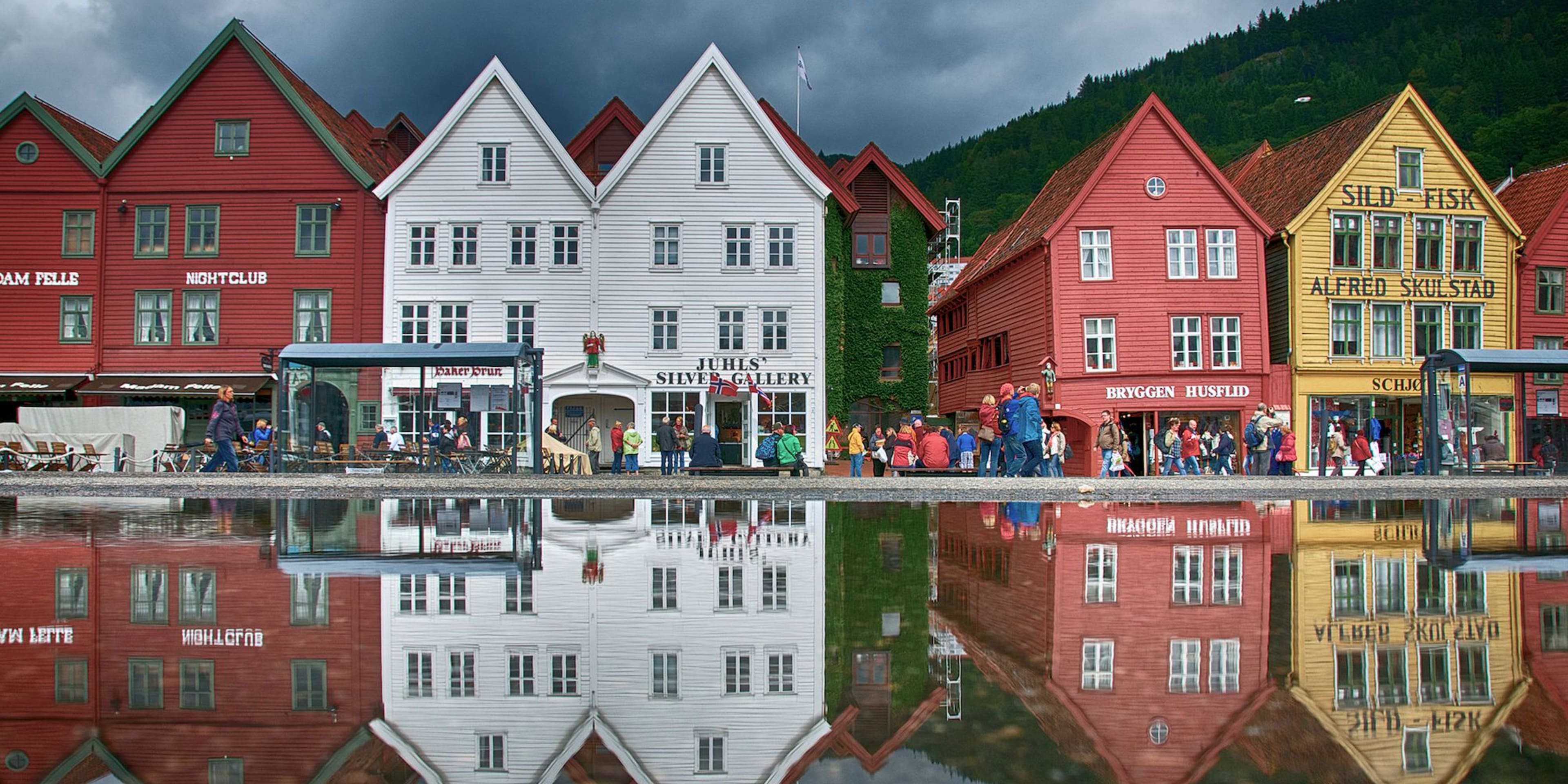 Fargerike Bryggen-bygninger i Bergen speiles i en hav, folk går under dramatiske skyer.