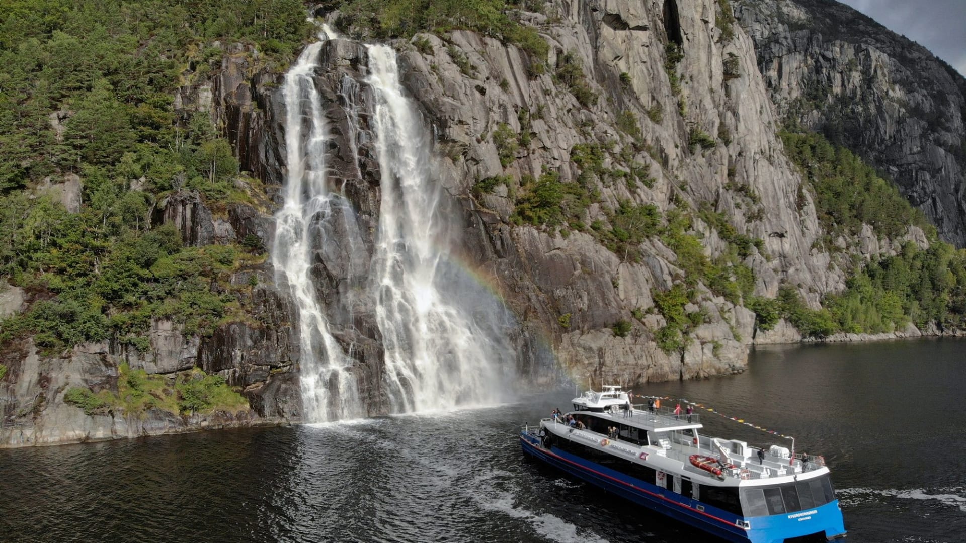 Tourboot fährt an Wasserfall in Lysefjord mit Regenbogen vorbei.