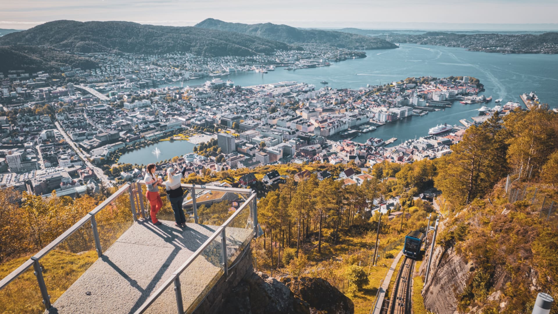 Zwei Personen stehen auf dem Aussichtspunkt am Fløyen mit Blick über Bergen, während die Fløibanen unterhalb den Hang hinauffährt.