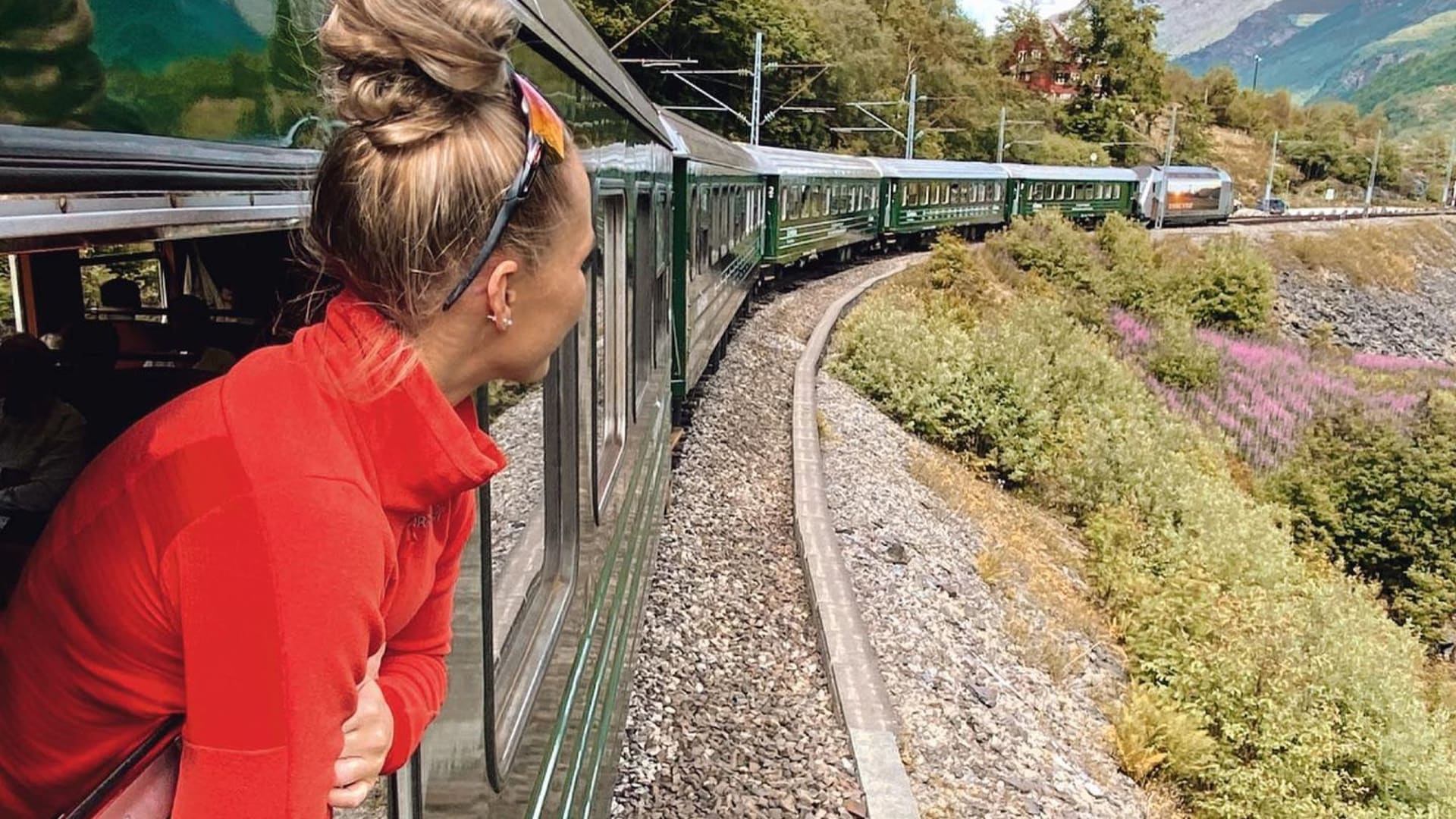Woman leans from Flåm railway  train, enjoying wildflowers and green mountain track.