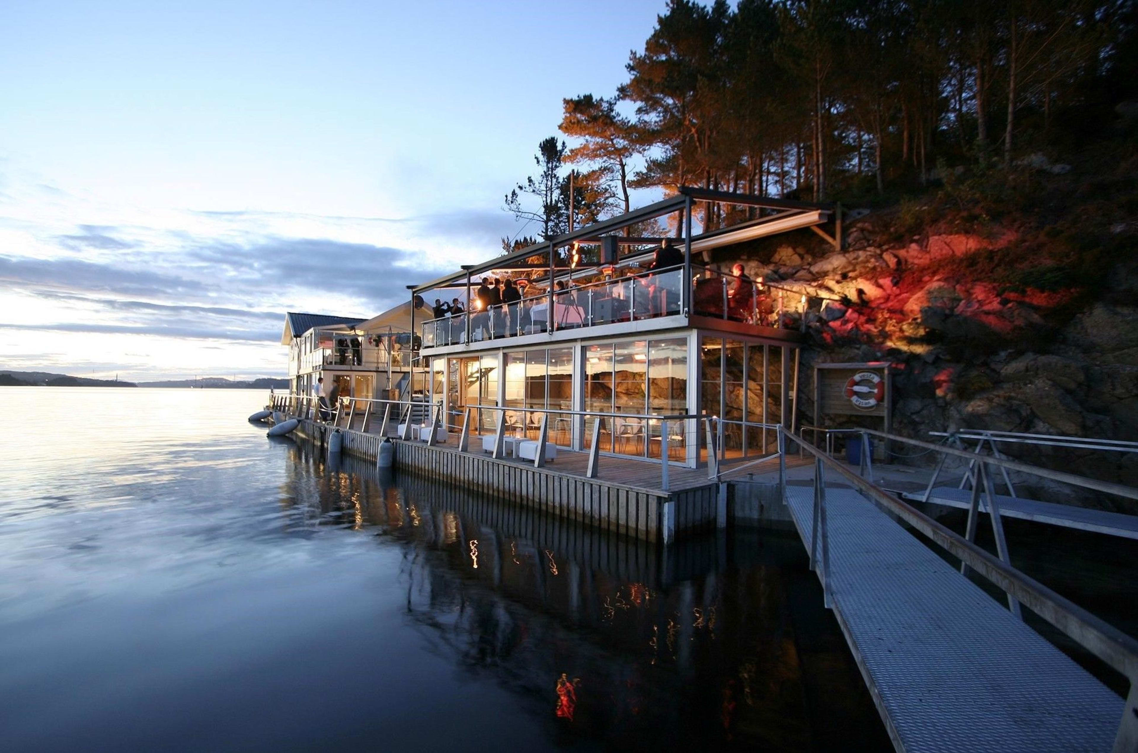Cozy glass-fronted waterfront restaurant glowing warmly at twilight, reflections shimmering on calm water surface.