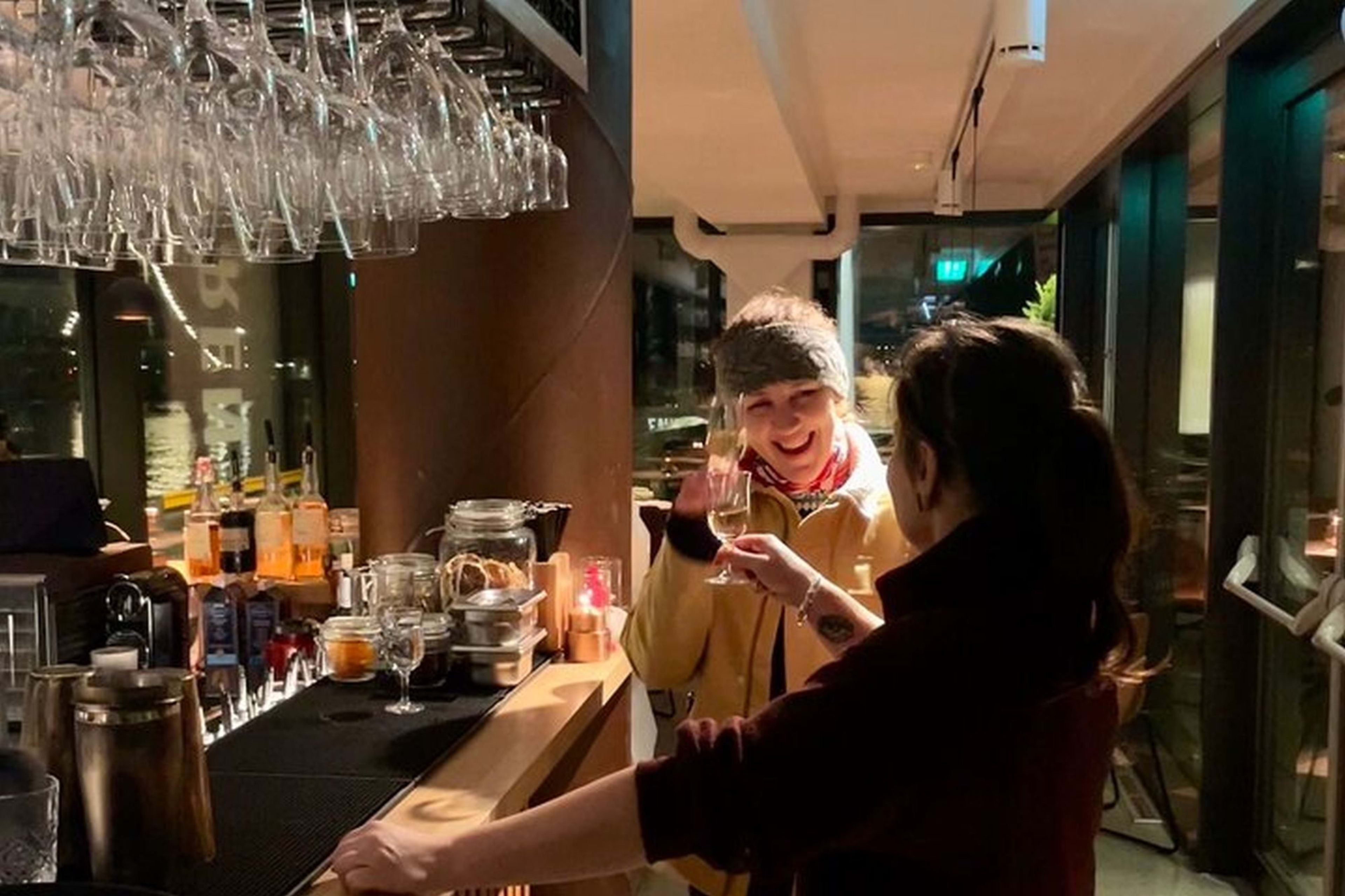 To kvinner som smiler og snakker ved en stilfull bar i Oslo med koselig belysning, champagne i hånden og utsikt over vannet.