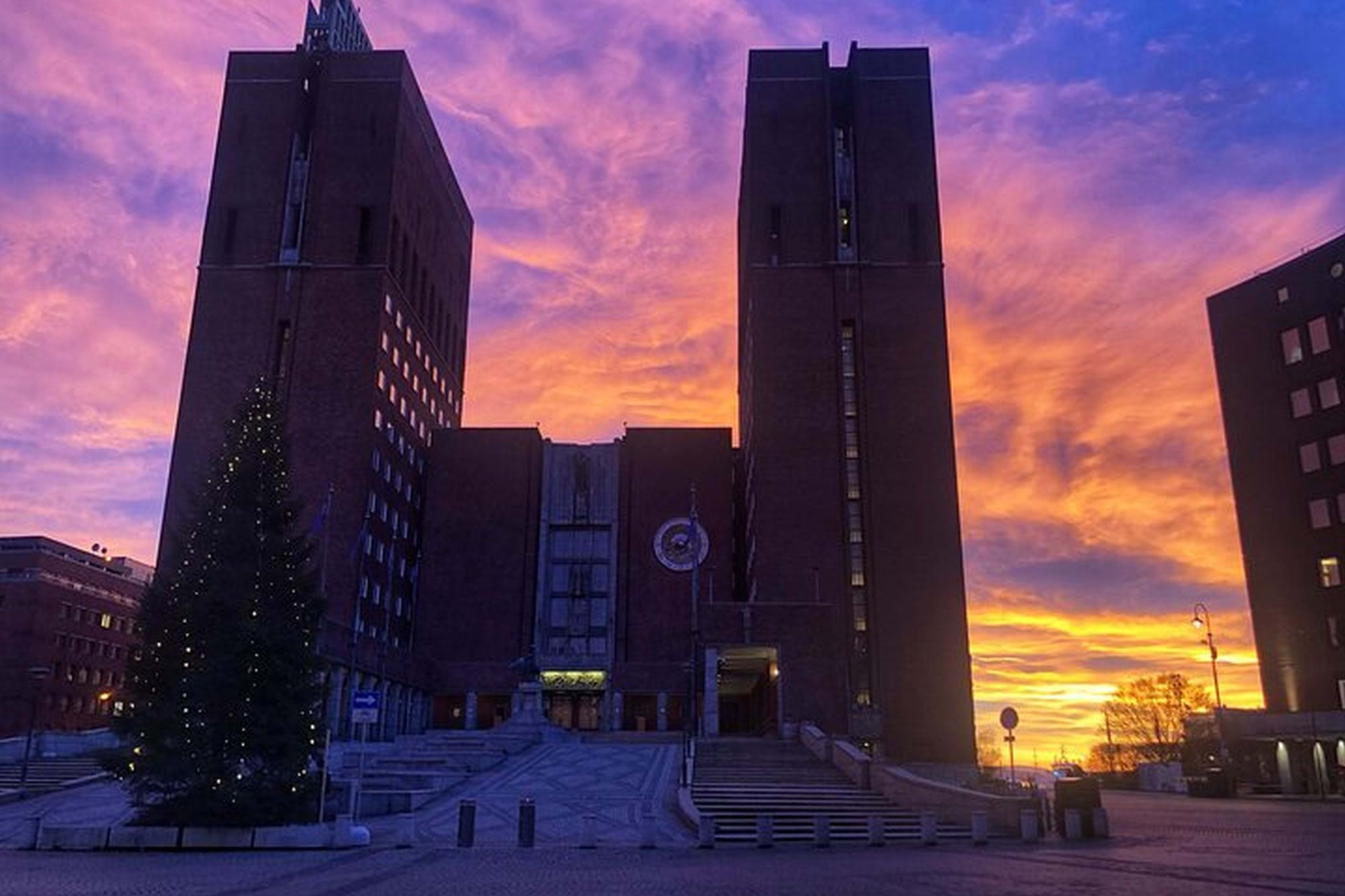 Dramatisk solnedgang med livlig lilla, rosa og oransje himmel over en symmetrisk murbygning, med et pyntet juletre.