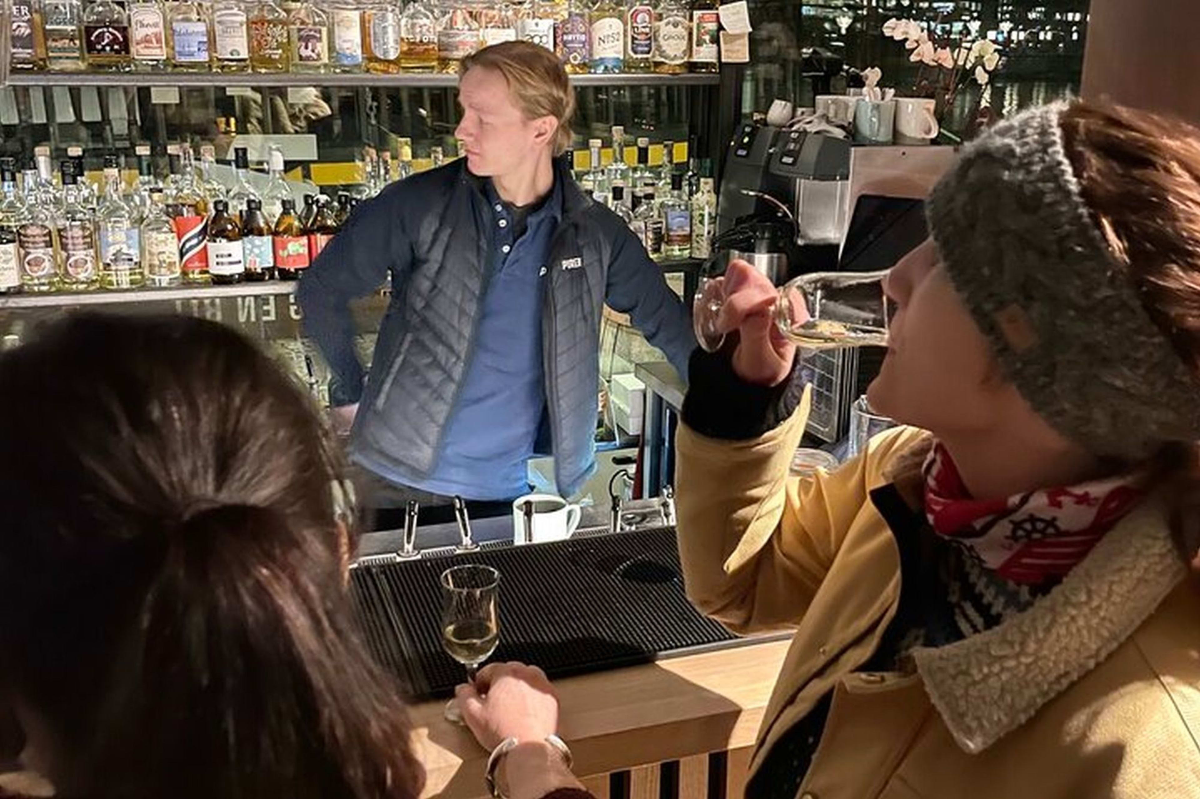 To kvinner på stilfull bar, den ene sipper på drinken mens den andre ser på, med bartender og hyller med flasker i koselig atmosfære.