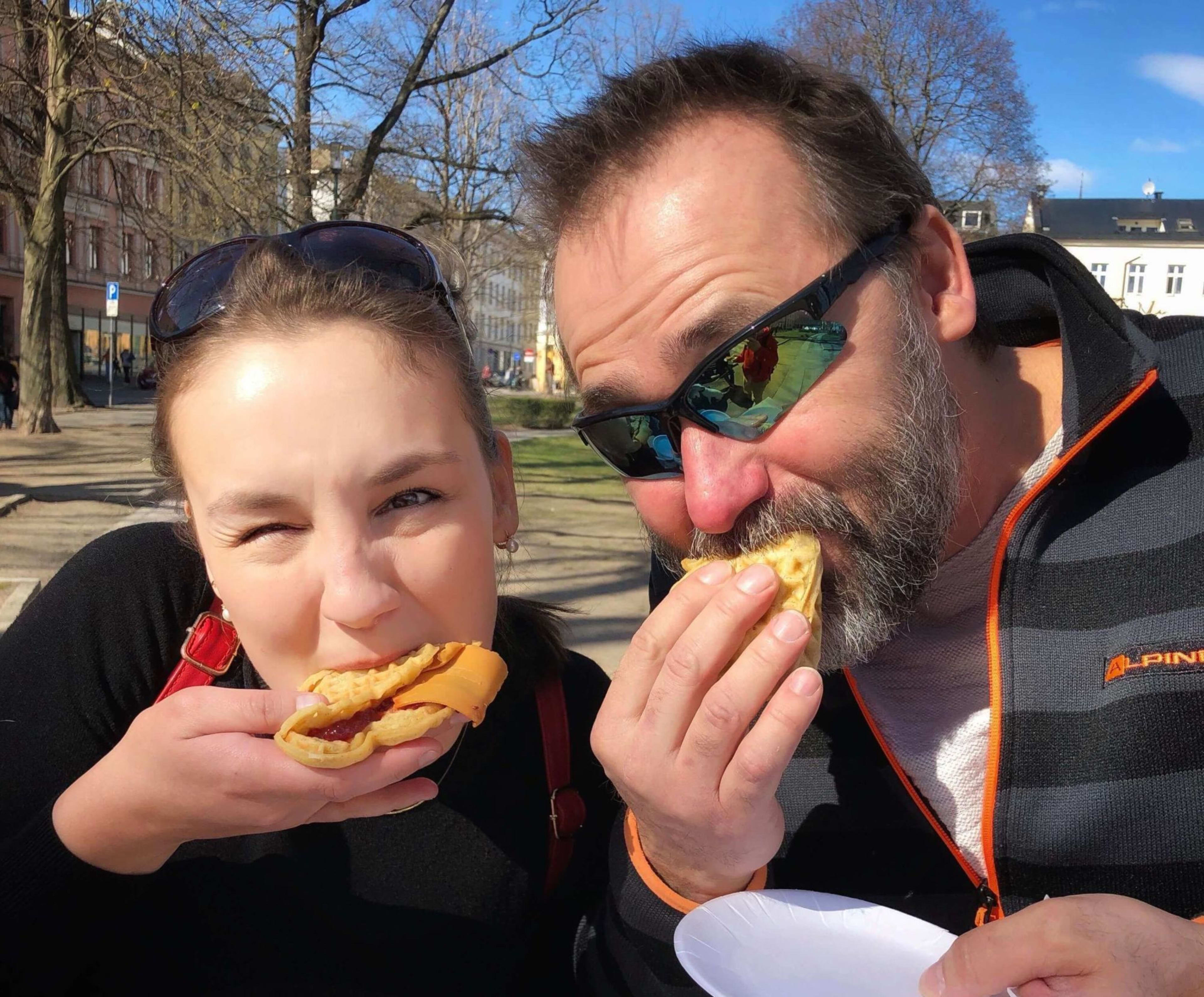 Glad duo som nyter vafler med ost og syltetøy i utendørs park, med solfylt vær og trær rundt.