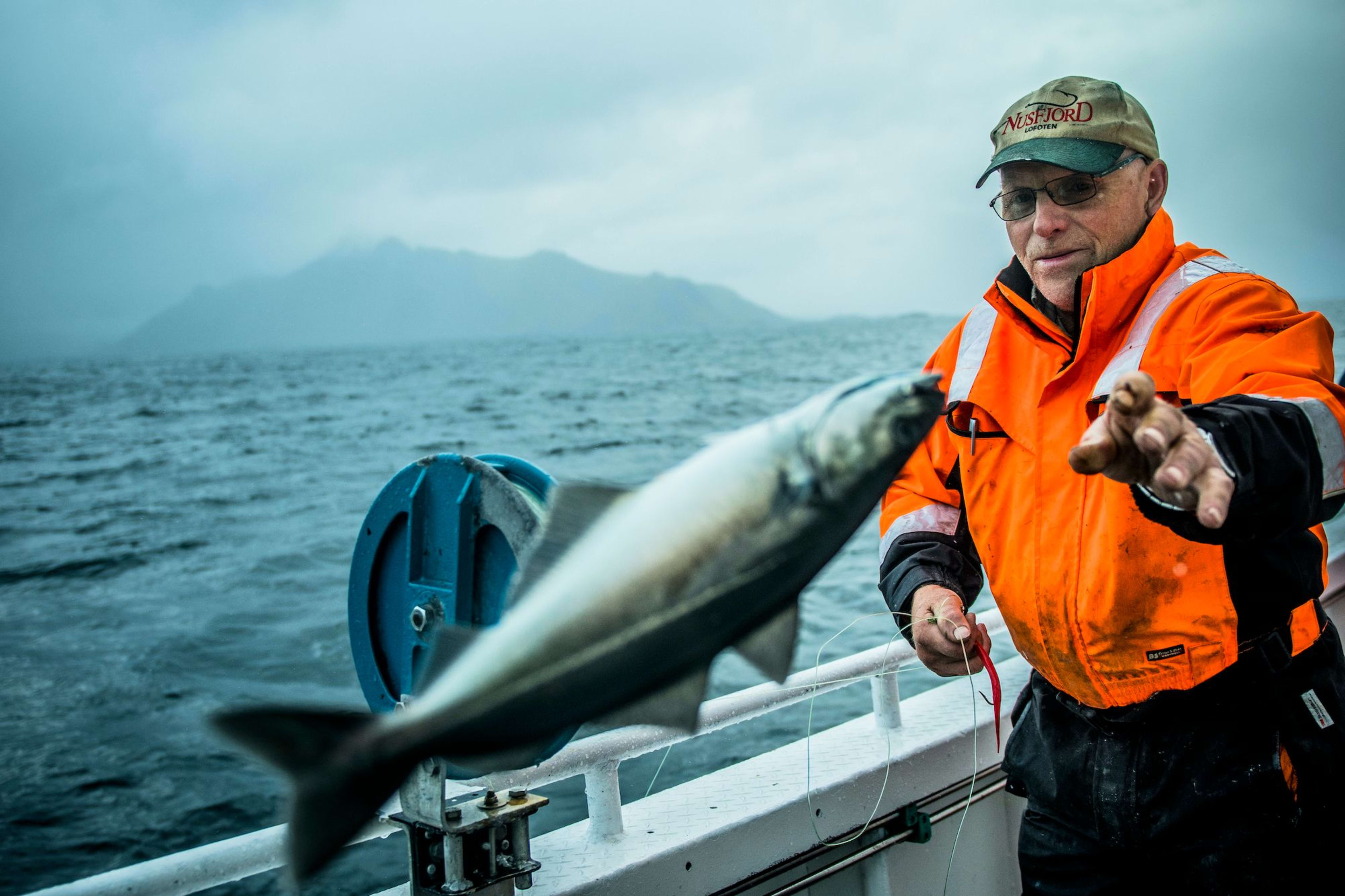 Mann i oransje jakke fanger fisk i lufta på sjøen.