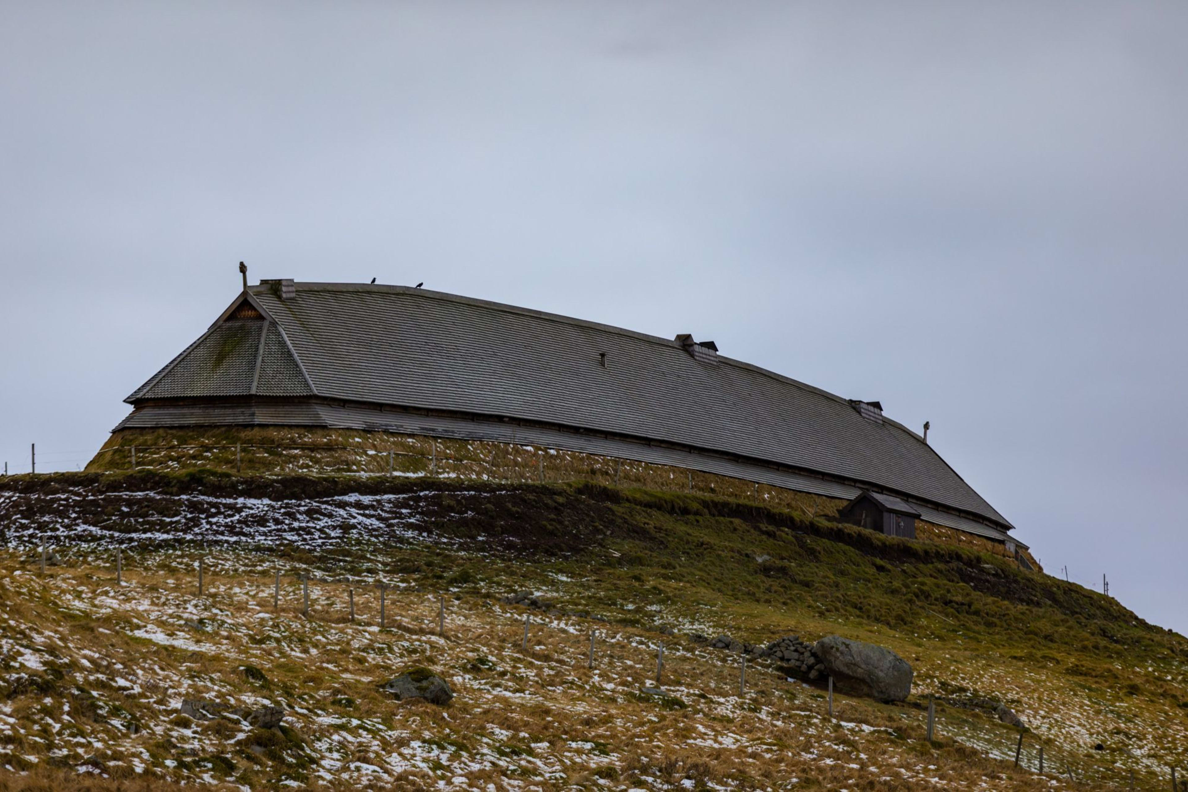 Viking-style longhouse on grassy hill with snow patches, wooden structure, overcast sky creating somber, cold atmosphere.