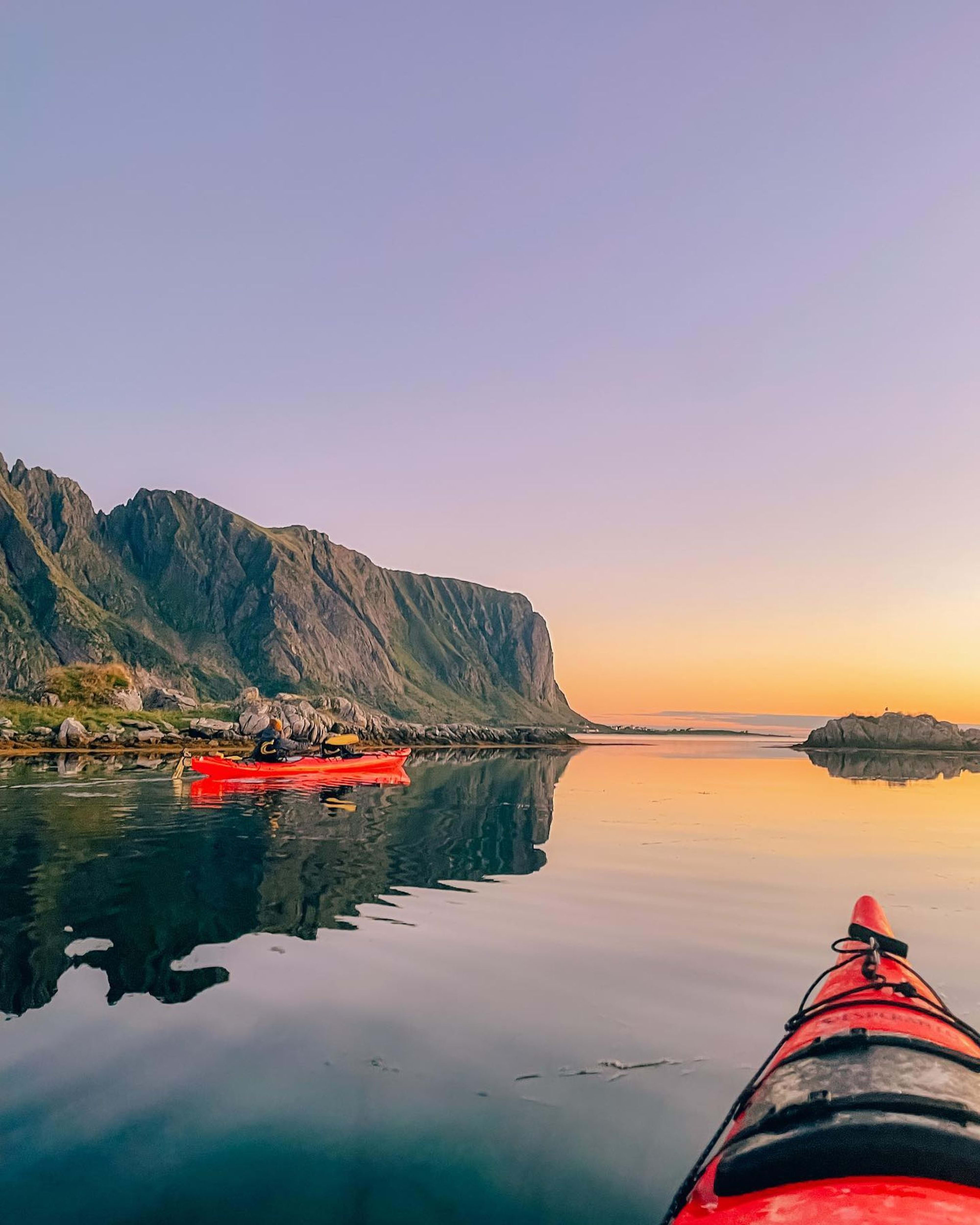 Zwei rote Kajaks gleiten bei Sonnenuntergang durch ruhigen Fjord mit Spiegelung.