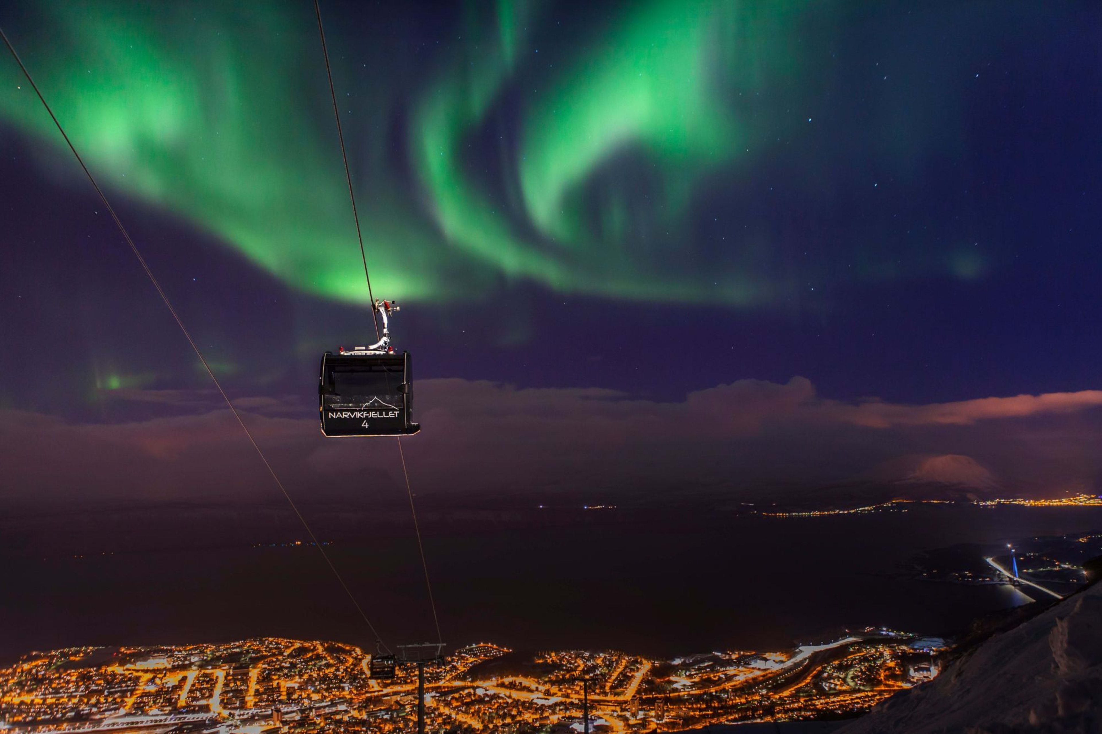 Narvik Seilbahn, die über eine Stadt aufsteigt, mit Nordlichtern, die den dunklen Himmel erleuchten, schneebedeckten Bergen und Stadtlichtern.
