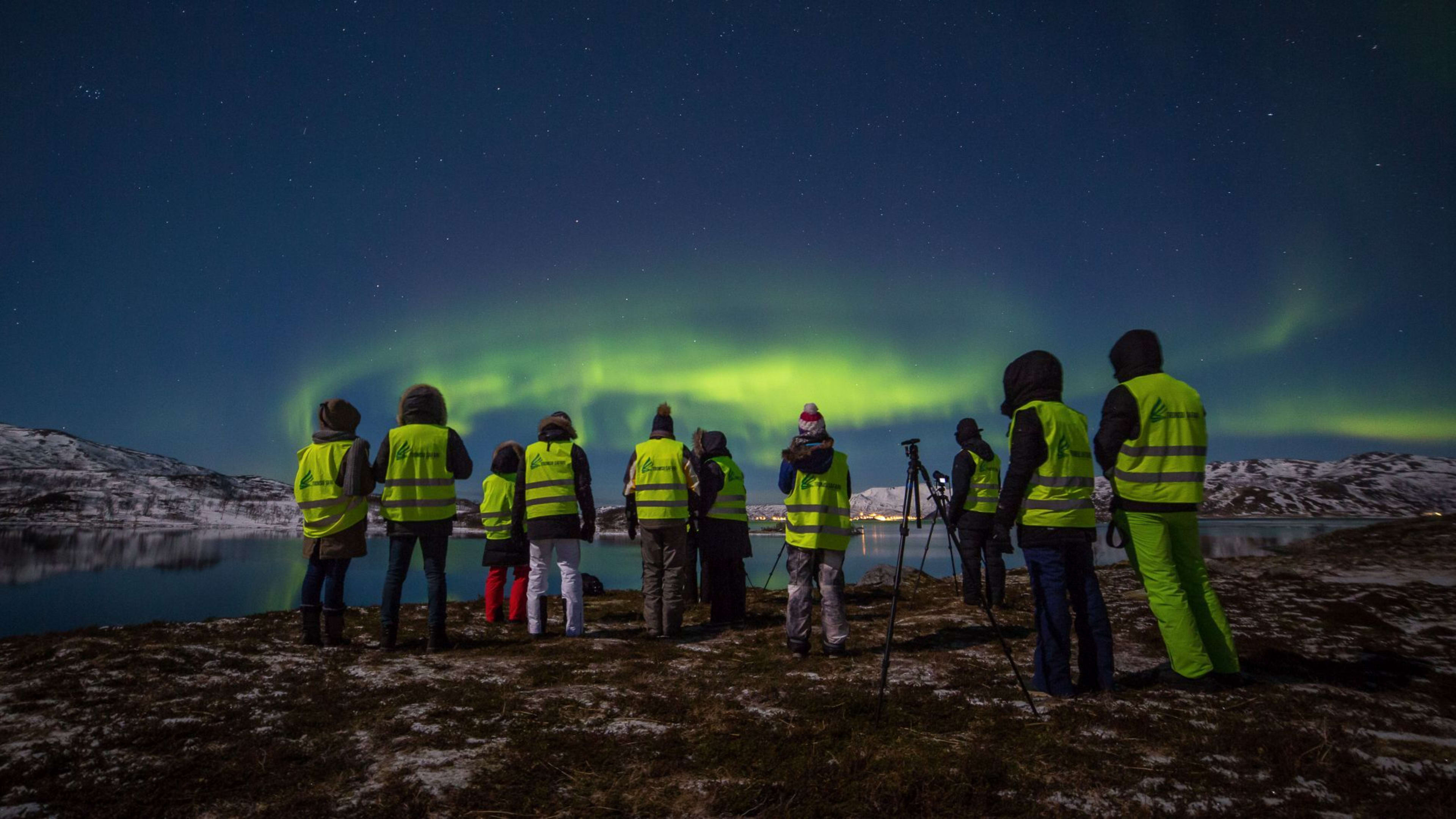 Folk i gule vester ser på nordlyset over et snødekt landskap med innsjø og fjell i bakgrunnen.