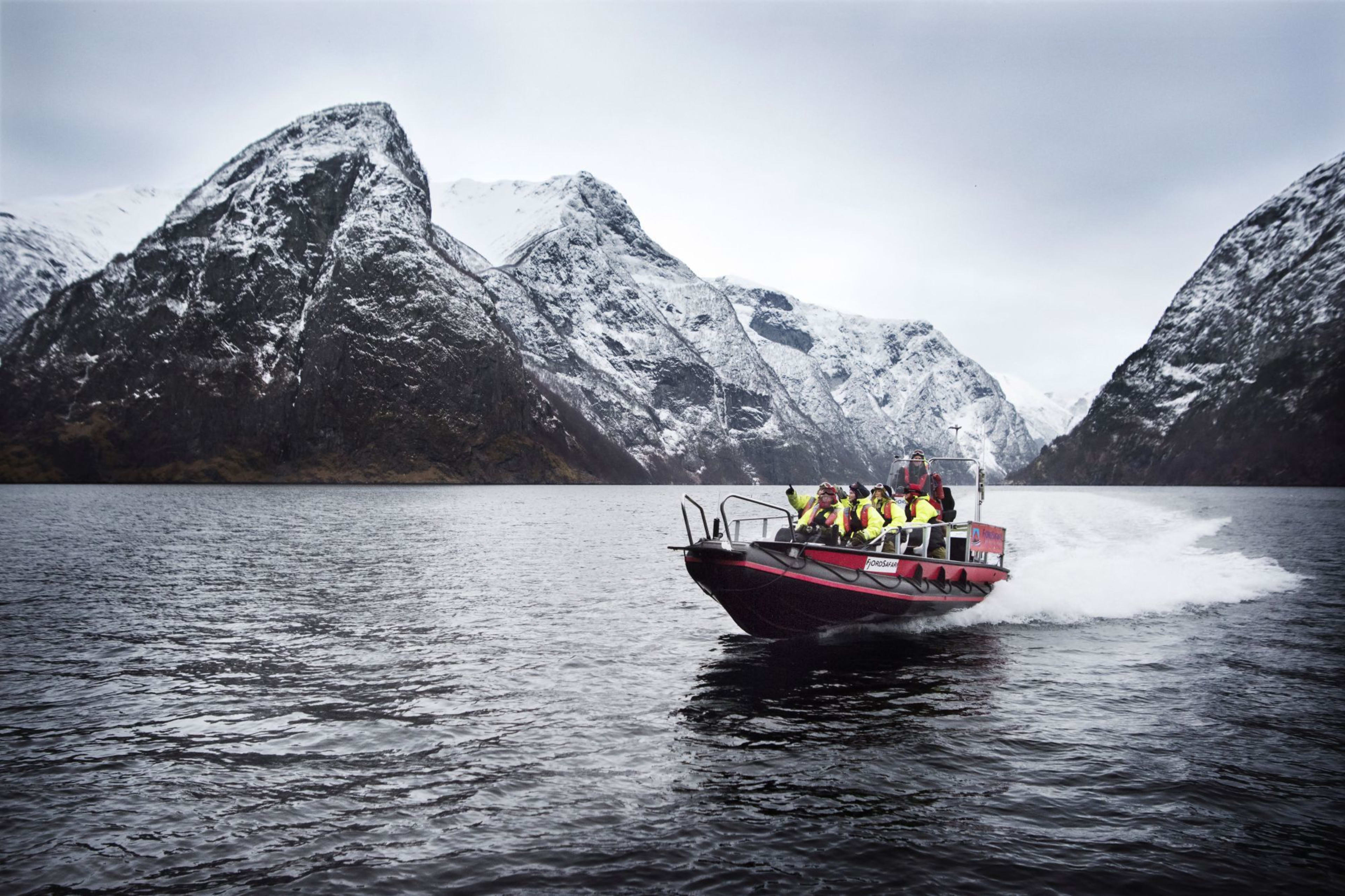 Rød oppblåsbar båt som raser gjennom ripplende vann, med personer i gule jakker, rammet av snøkledde fjell.