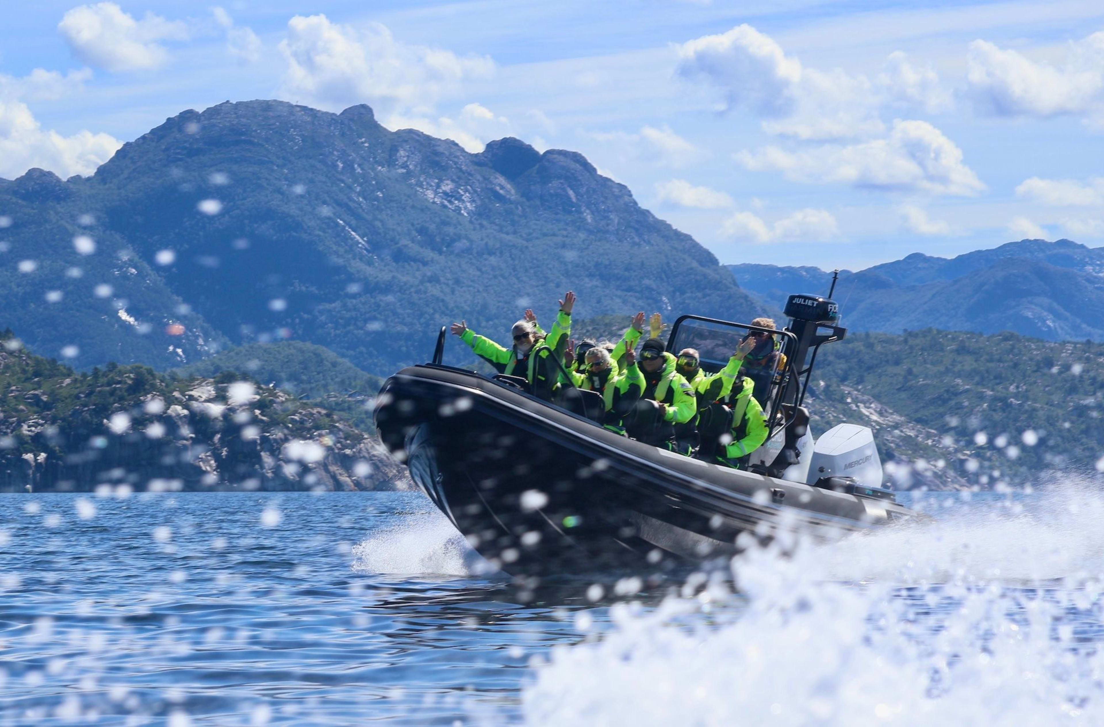 Ein schwarzes RIB-Boot mit Passagieren in neon-grünen Jacken, die mit Begeisterung die Arme heben, Berge steigen im Hintergrund auf.