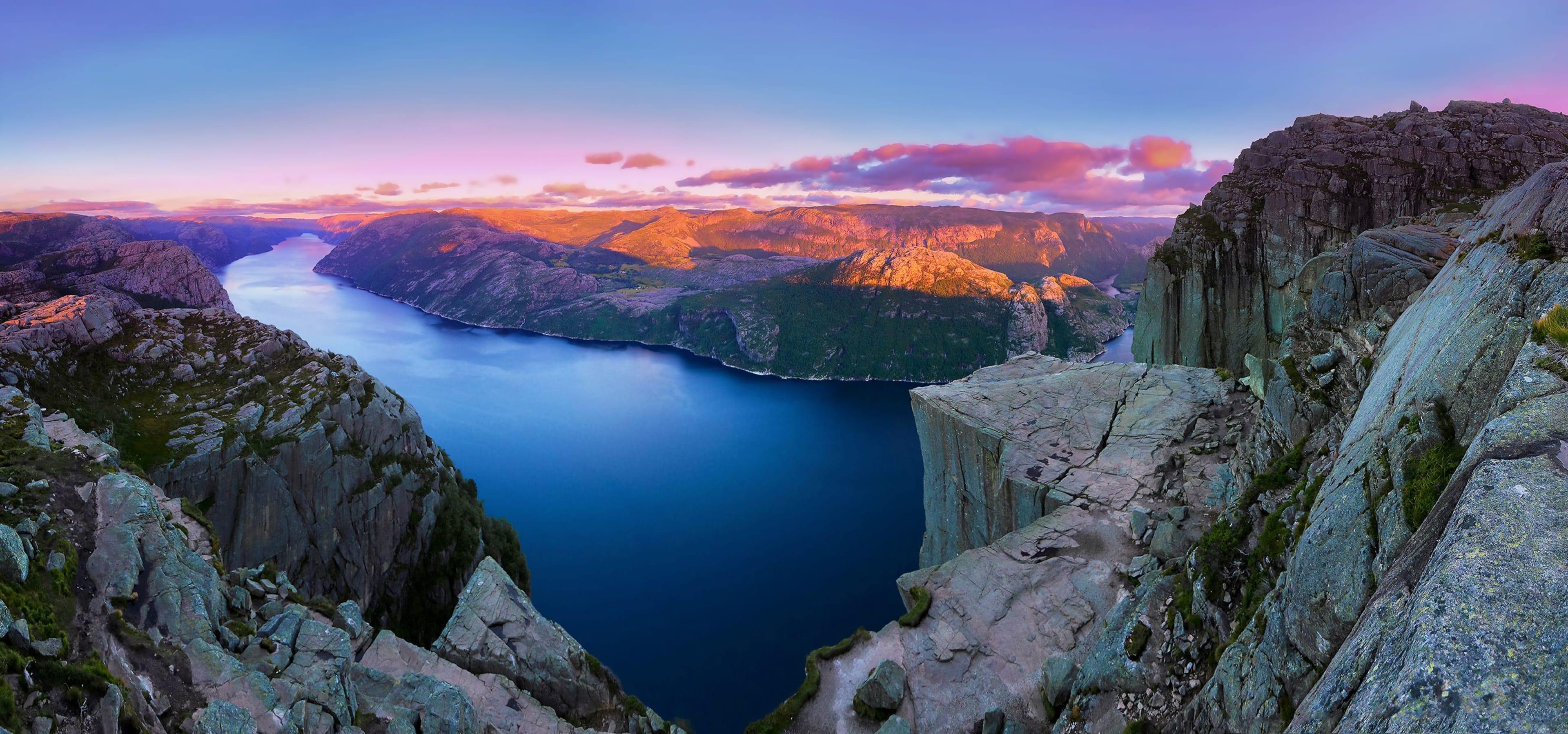 Preikestolen-Blick auf den Lysefjord bei Sonnenuntergang mit warmem Licht auf schroffen Gipfeln unter einem farbenfrohen Himmel.