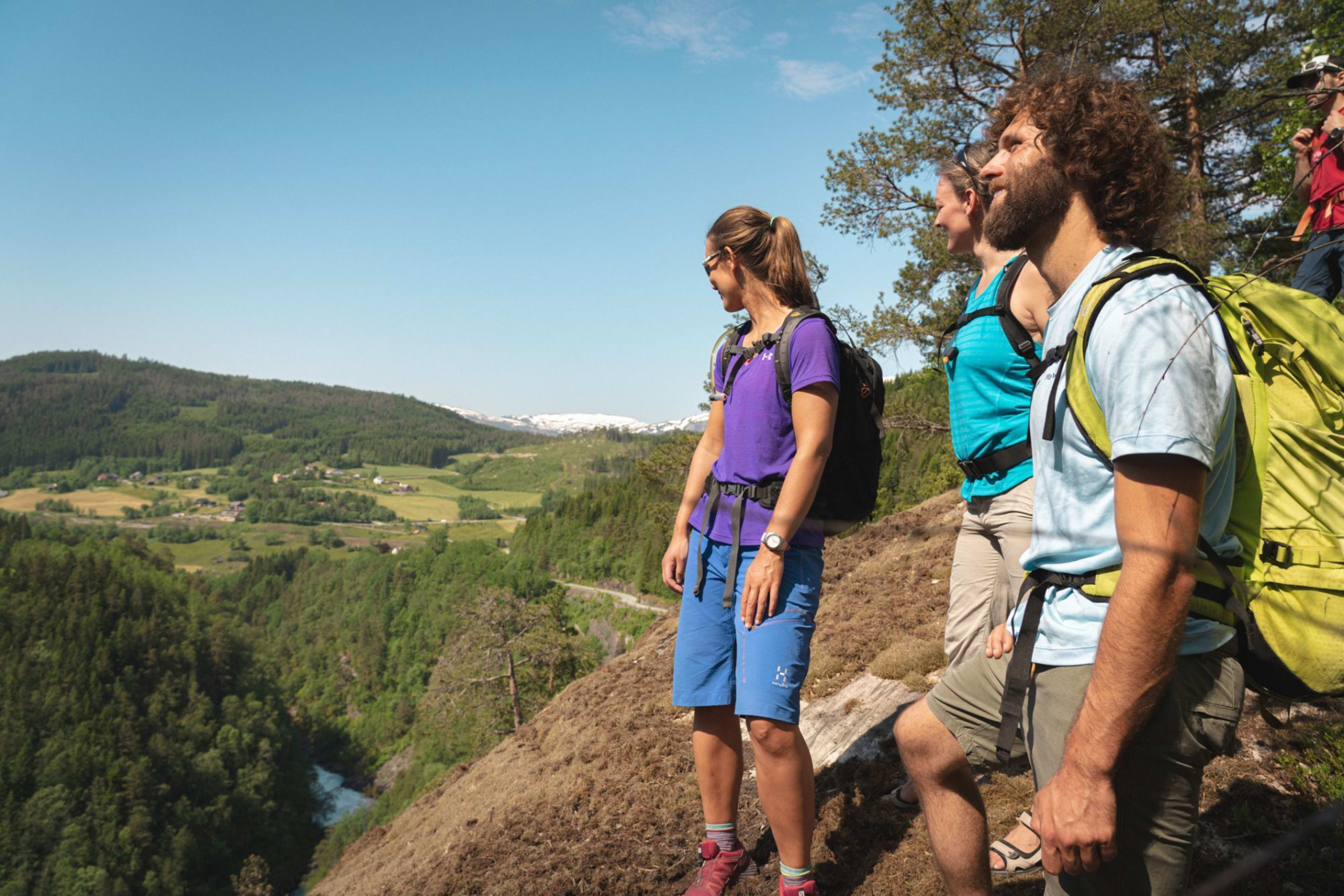 Group of hikers resting on grassy hillside, smiling and admiring panoramic view of valley with farms and roads.
