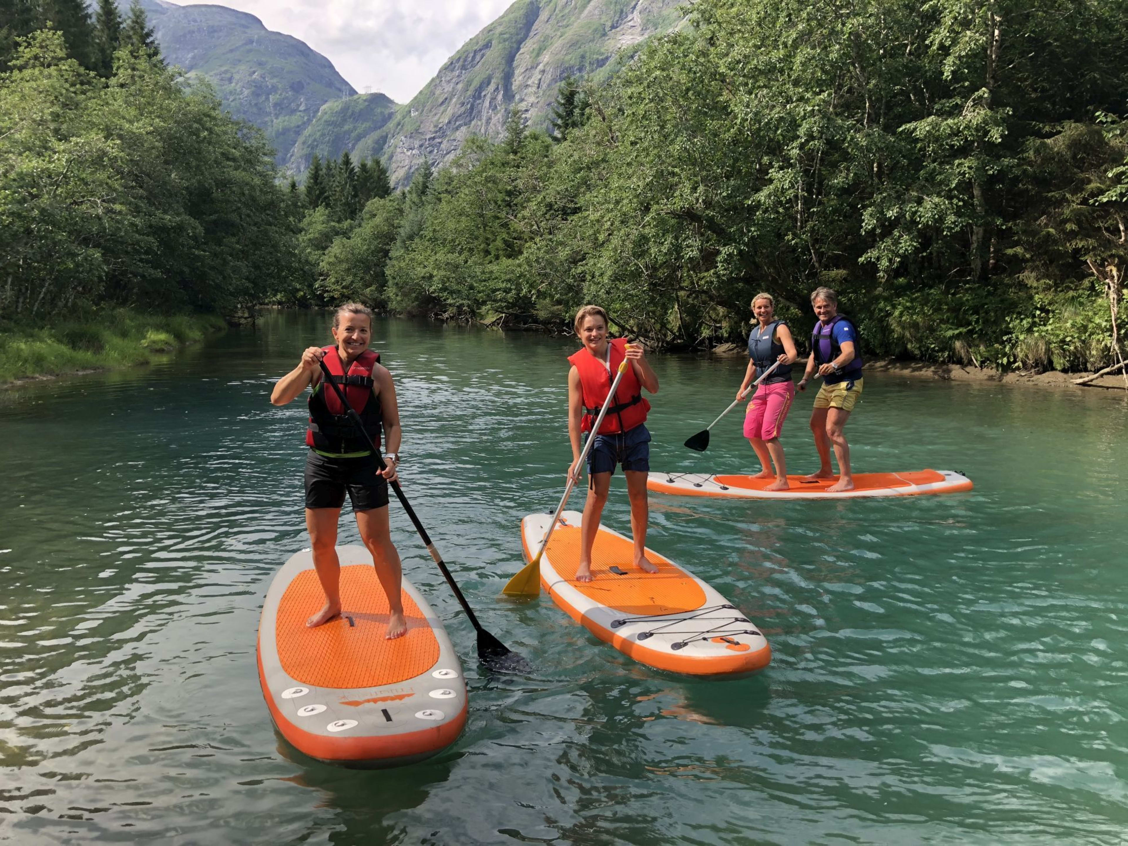 Vier Personen stehen auf Stand-Up-Paddleboards auf einem kristallklaren Fluss, umgeben von üppigem Grün und Bergen.