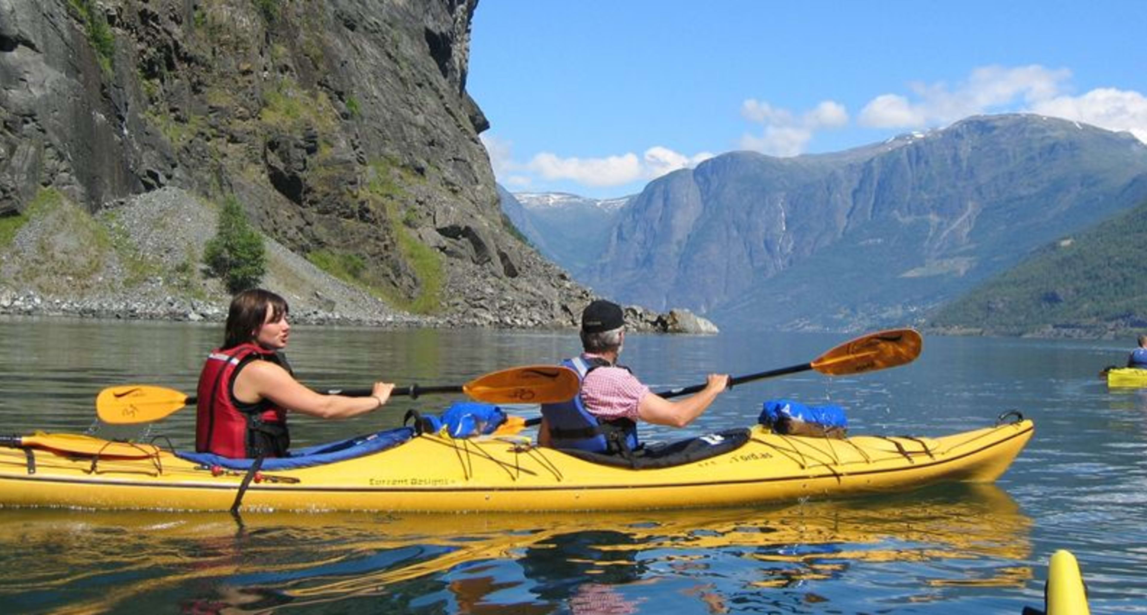 To kajakkpadlere i en gul tandemkajakk padler på klart vann med en fjellbakgrunn.