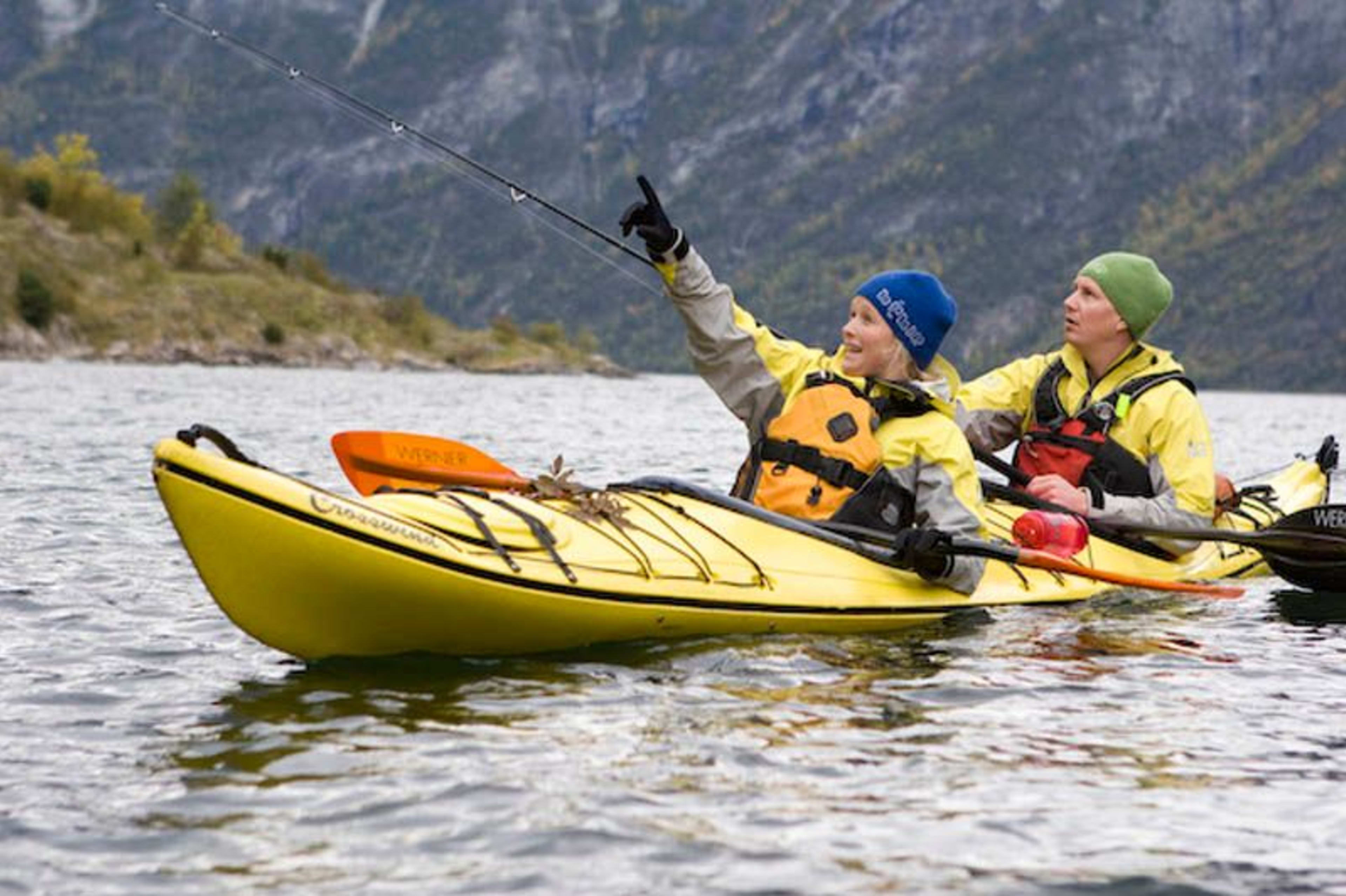 Zwei Kajakfahrer in einem gelben Tandemkajak paddeln auf einem ruhigen Fjord, wobei einer enthusiastisch auf etwas in der Ferne zeigt, umgeben von felsigen Klippen und Herbstlaub.