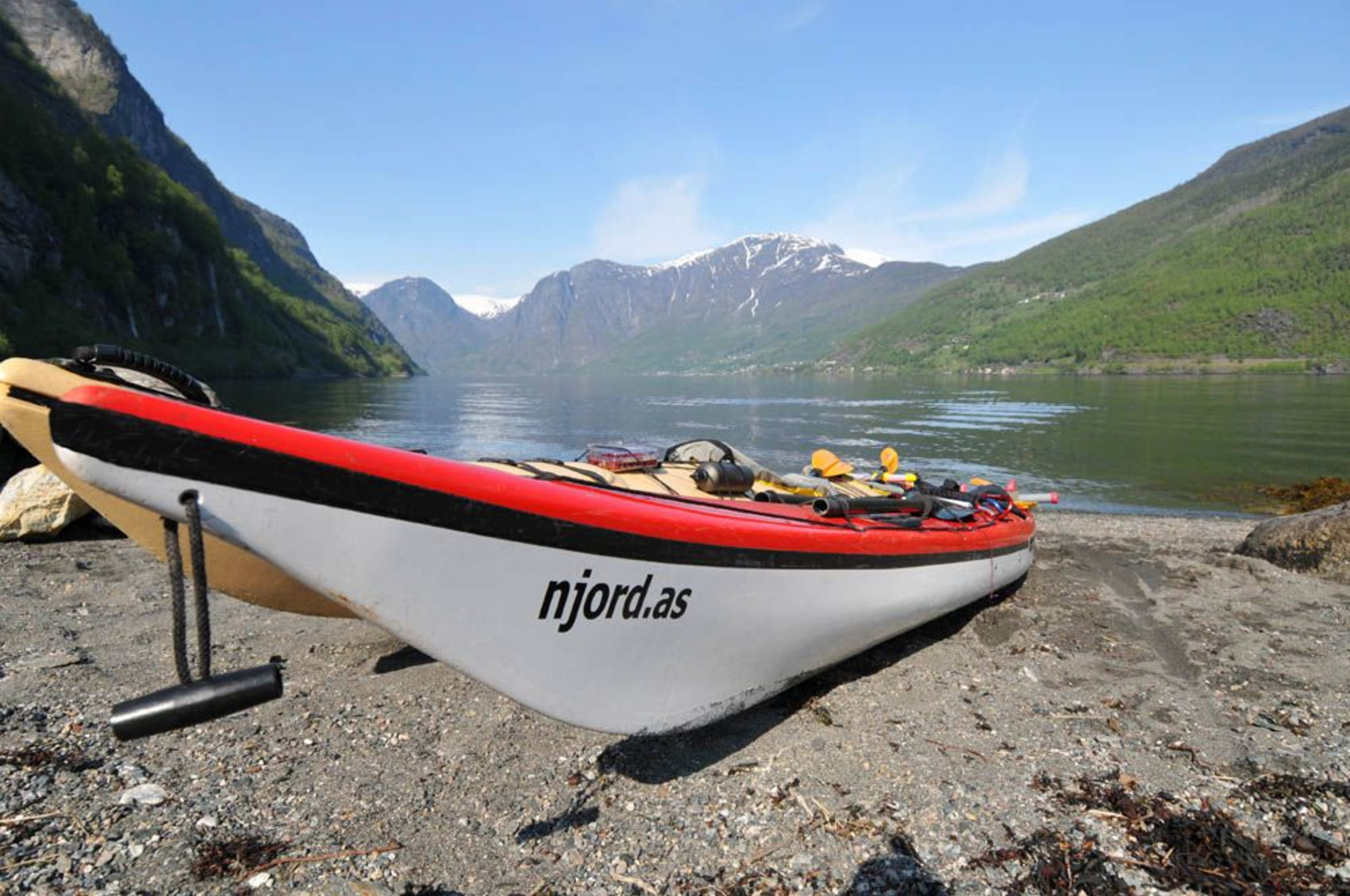 Hvit og rød kajakk merket "njord.as" hvilende på en sandstrand, klar for en fjordopplevelse med fjell og reflekterende vann i bakgrunnen.