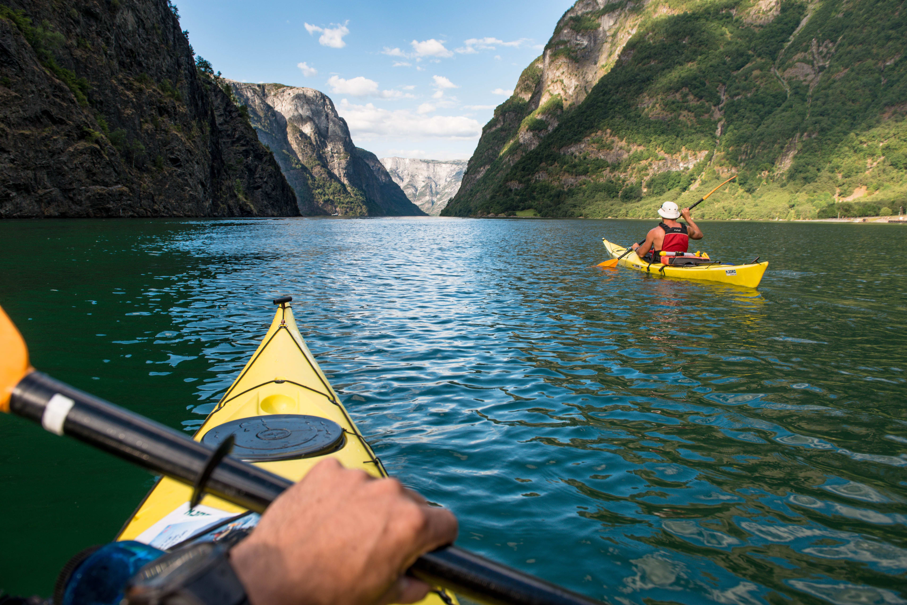 Kajakkpadler som padler en gul kajakk på en rolig, turkis fjord, omringet av tårnende klipper med frodig grønt landskap, tatt fra perspektivet til en annen kajakkpadler.