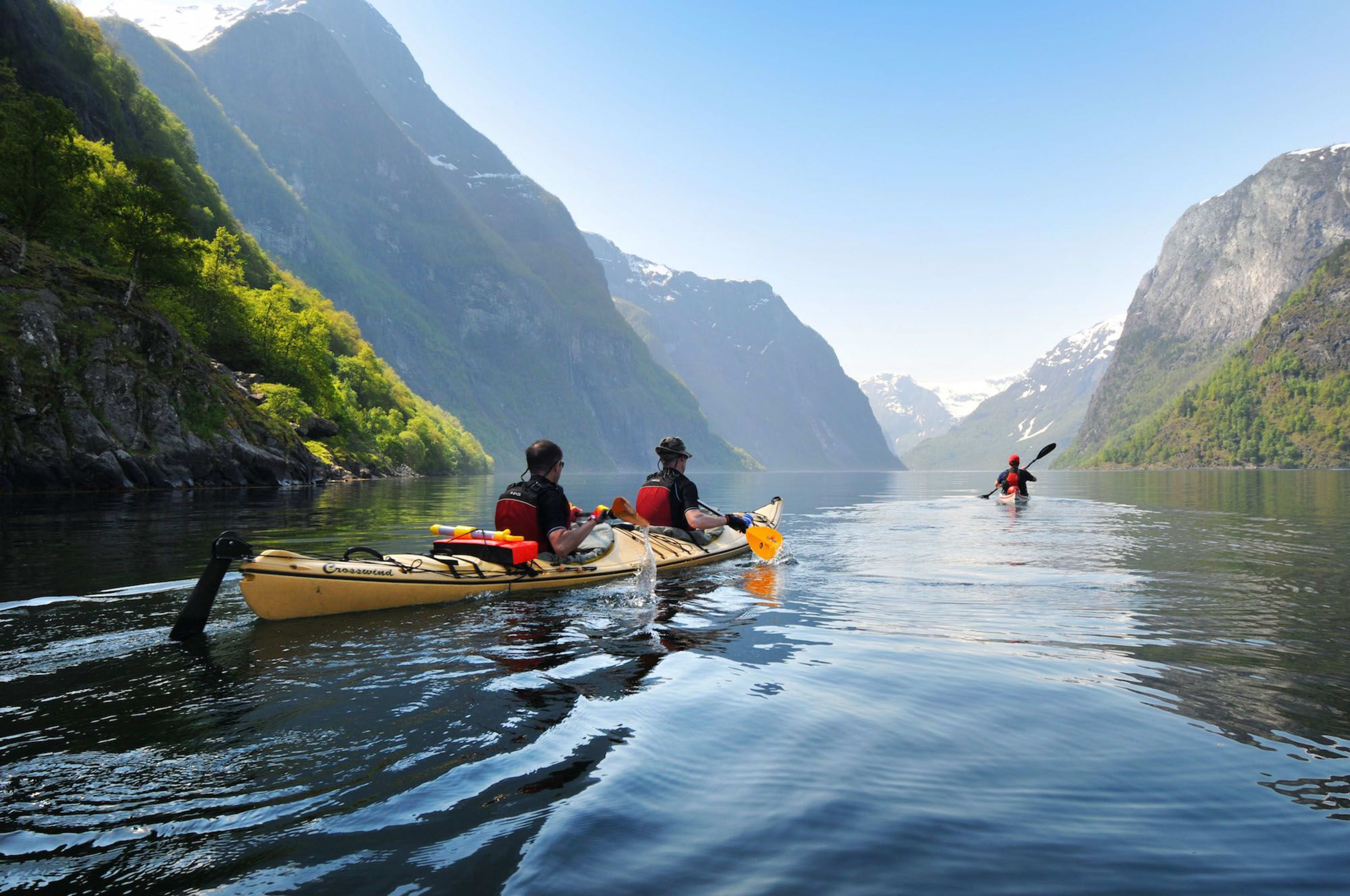 To kajakkpadlere padler langs en rolig, reflekterende fjord, omringet av tårnende grønne fjell og dramatiske klipper under en klar blå himmel.