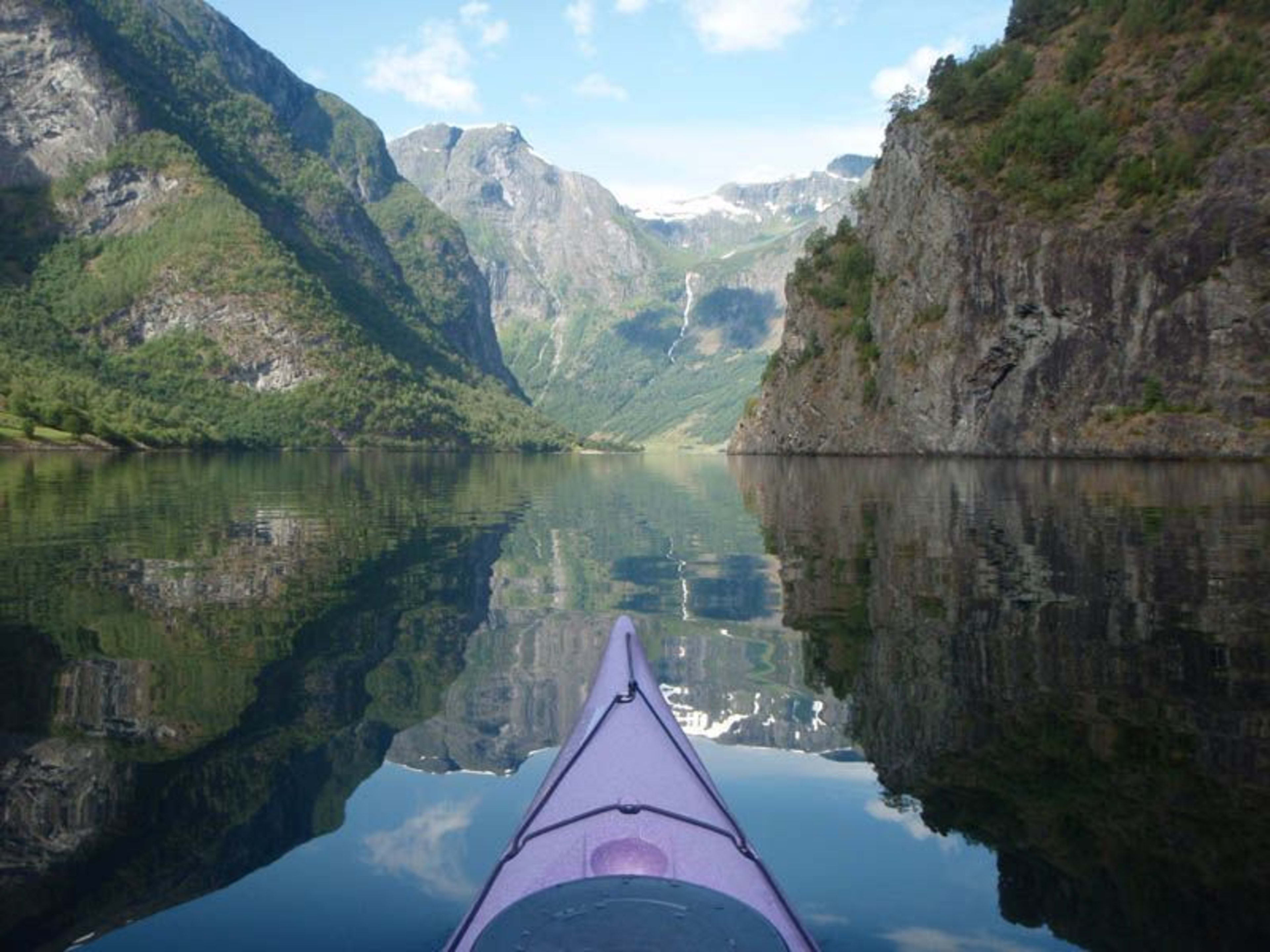 Roelig perspektiv fra en lilla kajakk som glir på rolig vann, omringet av tårnende klipper og fossende vannfall under en blå himmel.