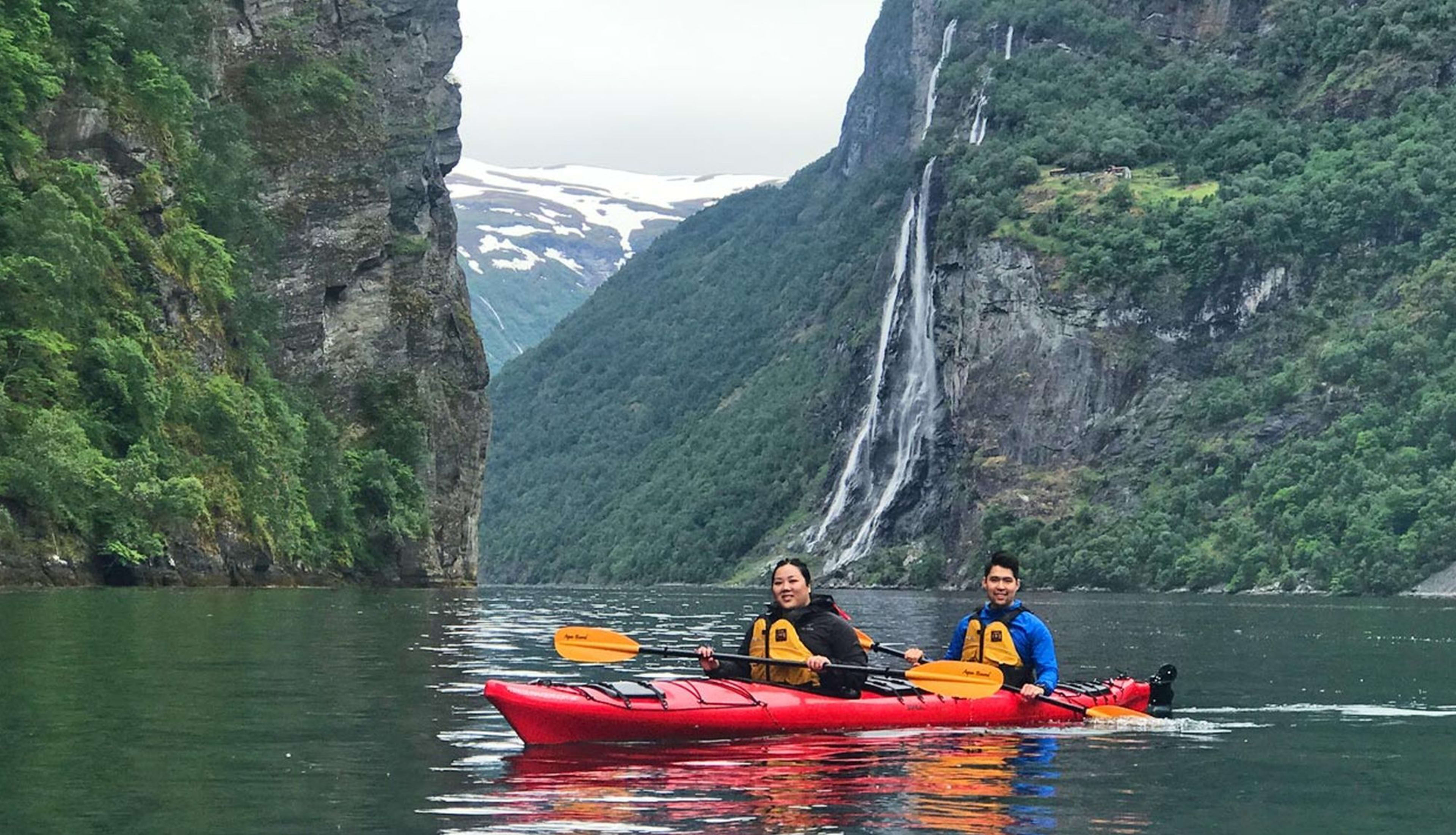 Zwei Kajakfahrer paddeln zusammen in einem roten Kajak durch einen norwegischen Fjord mit einem Wasserfall und schneebedeckten Bergen.