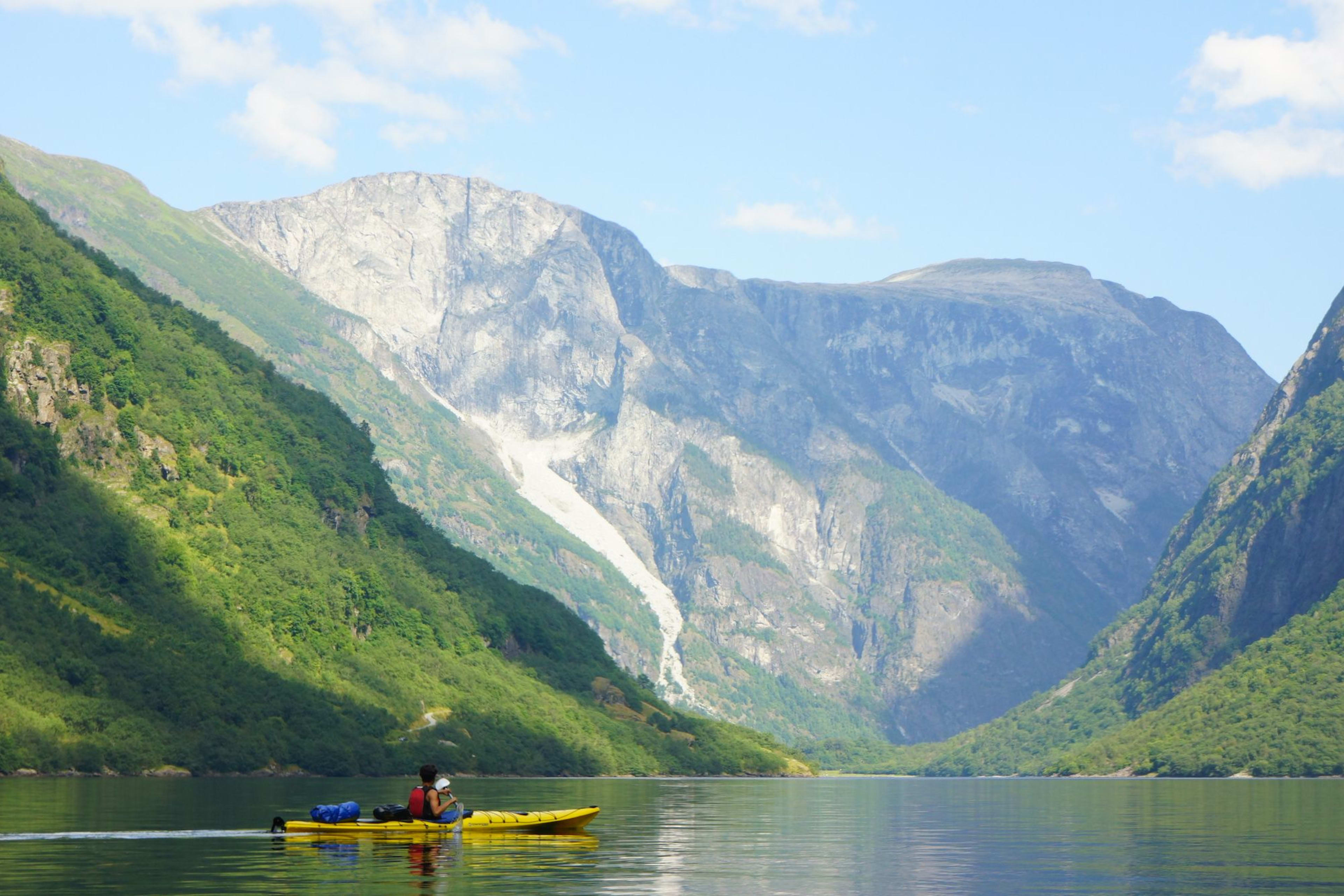 Person kayaking in a yellow kayak on calm water, surrounded by green hills and mountain cliffs, with a waterfall in the background.