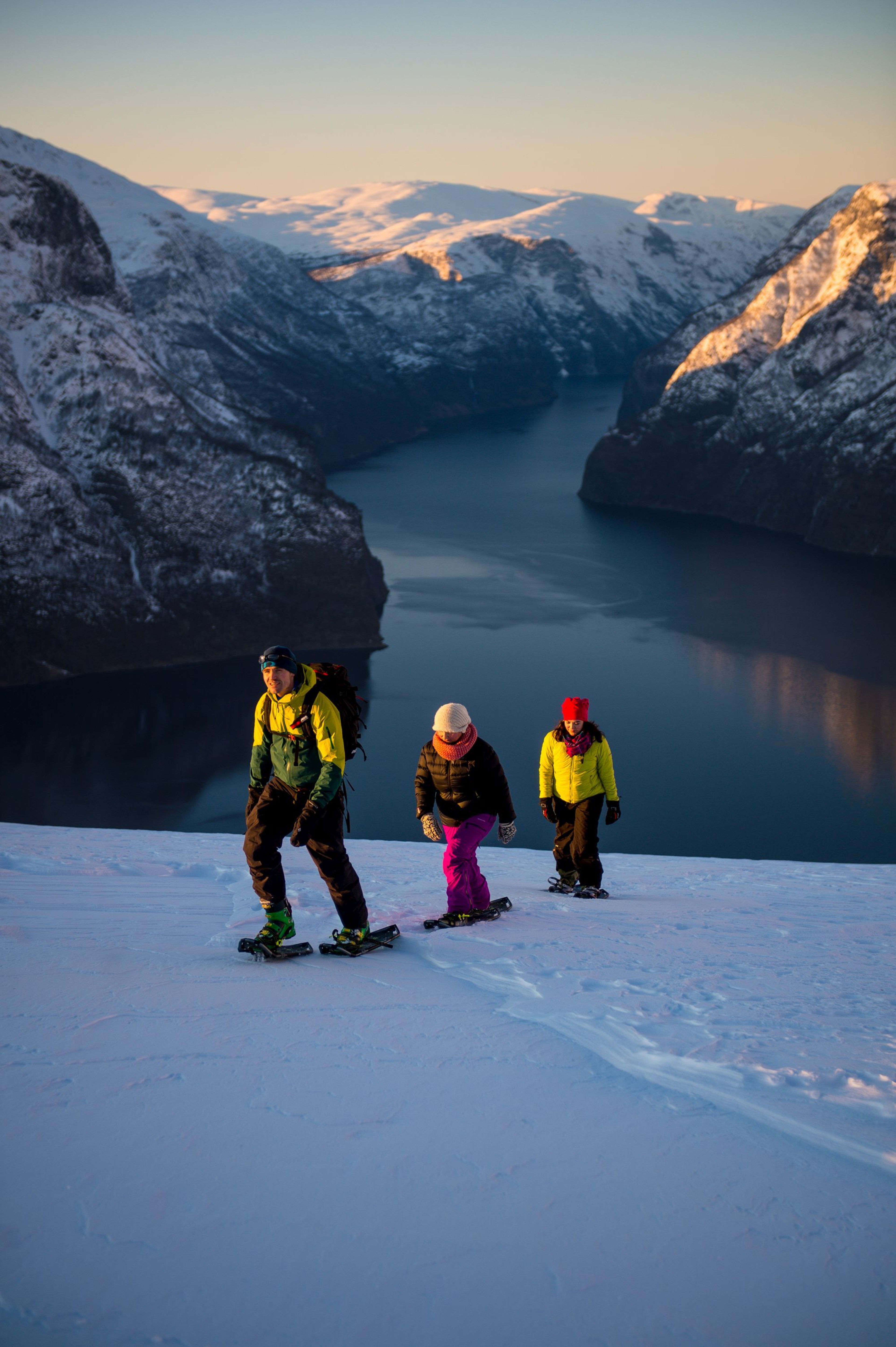 Three people snowshoe uphill at sunset with fjord and snowy peaks behind.