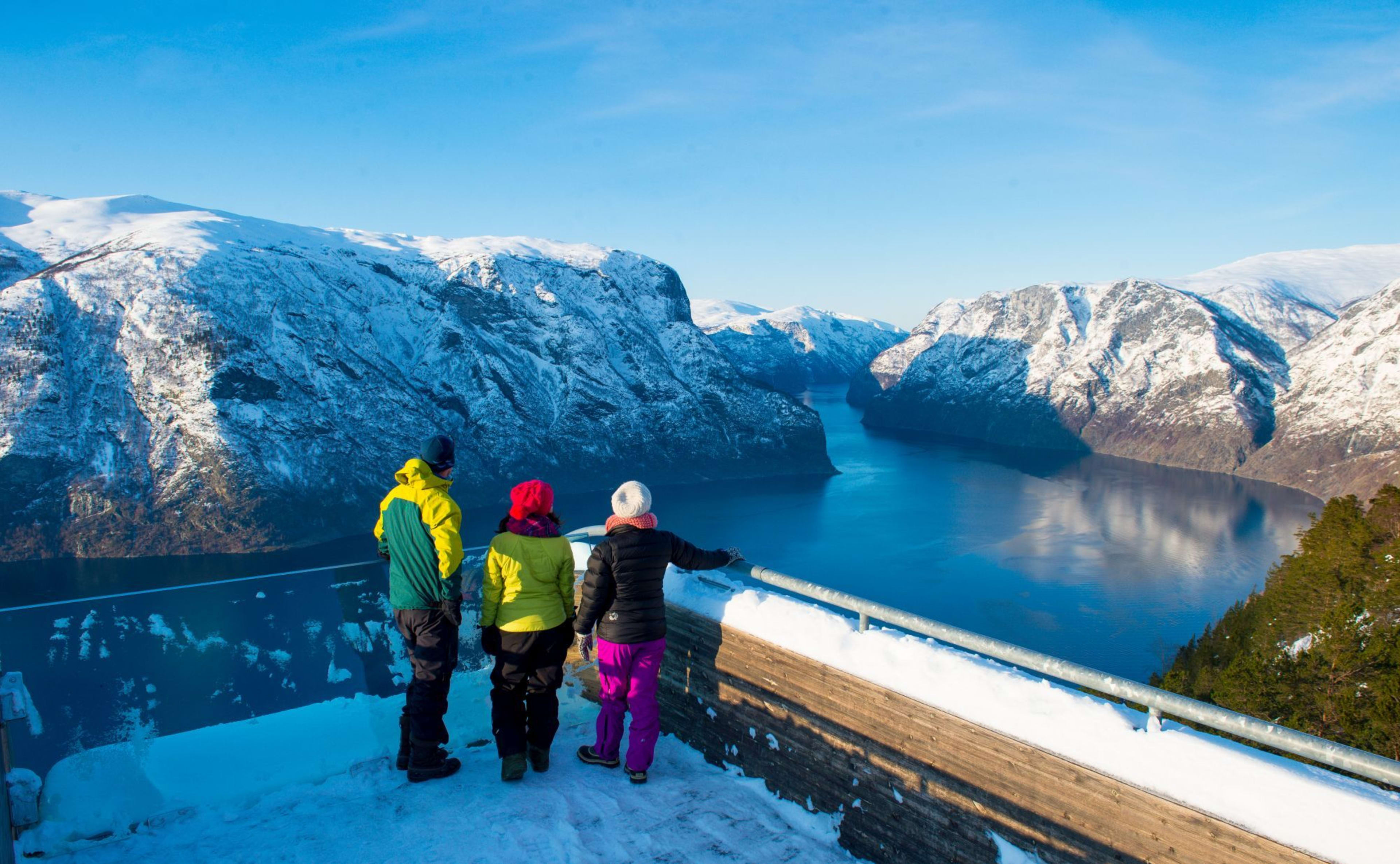 Three people on snowy viewpoint overlook blue fjord and snowy peaks.