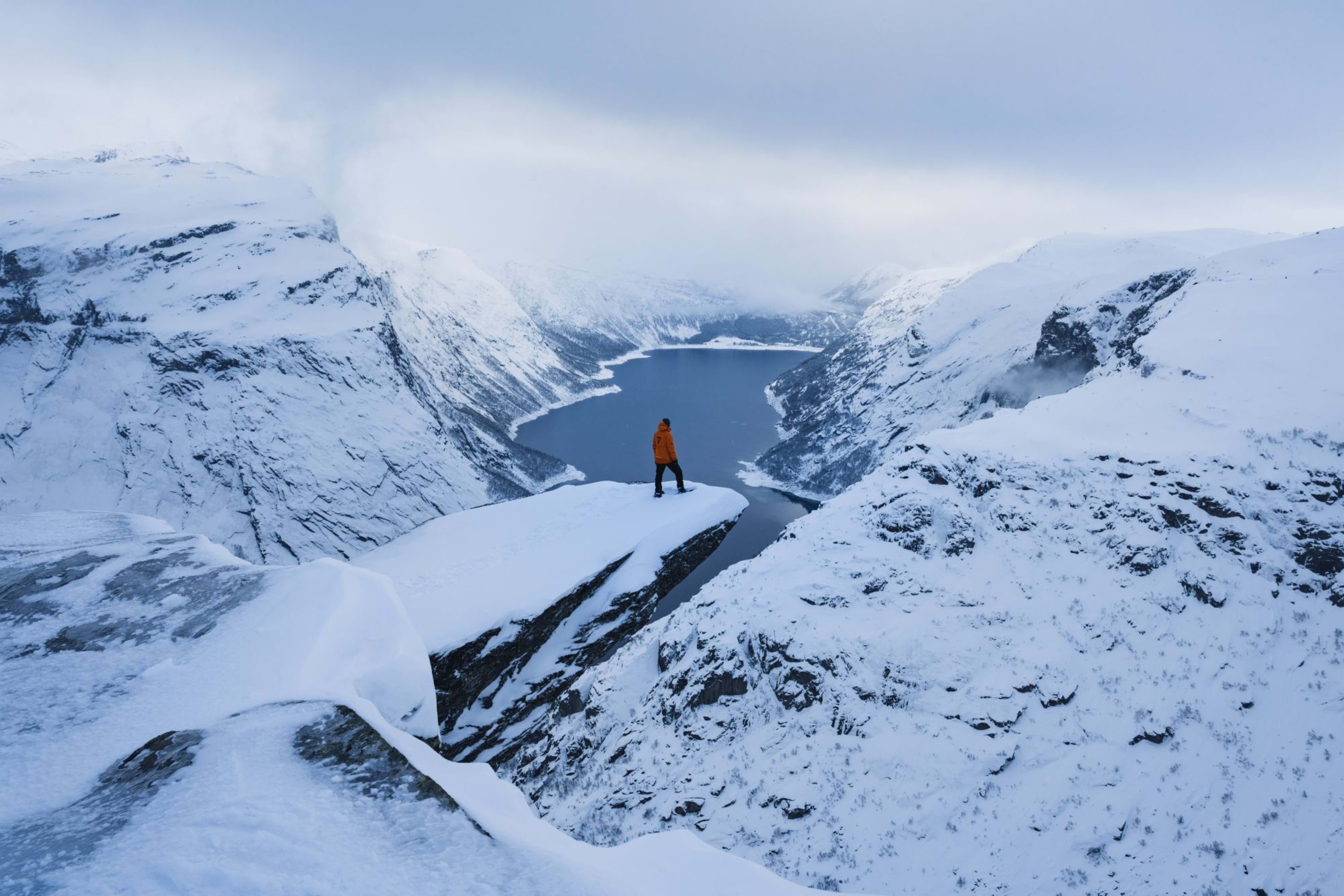 A person in an orange jacket stands alone on a cliff, looking over a fjord surrounded by snow-capped mountains.