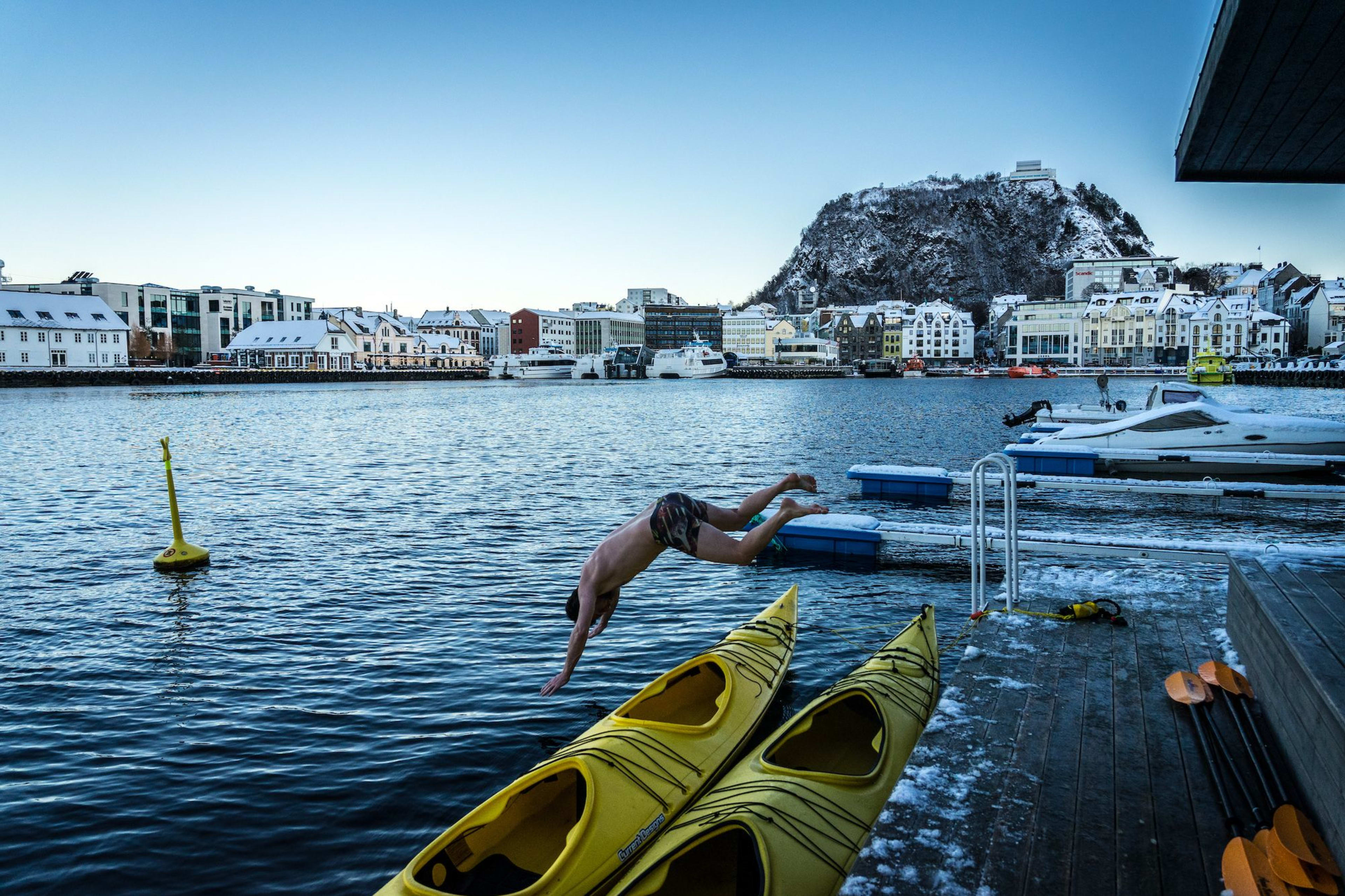 A person diving into cold water from a dock, with snowy buildings and a mountain in the background.