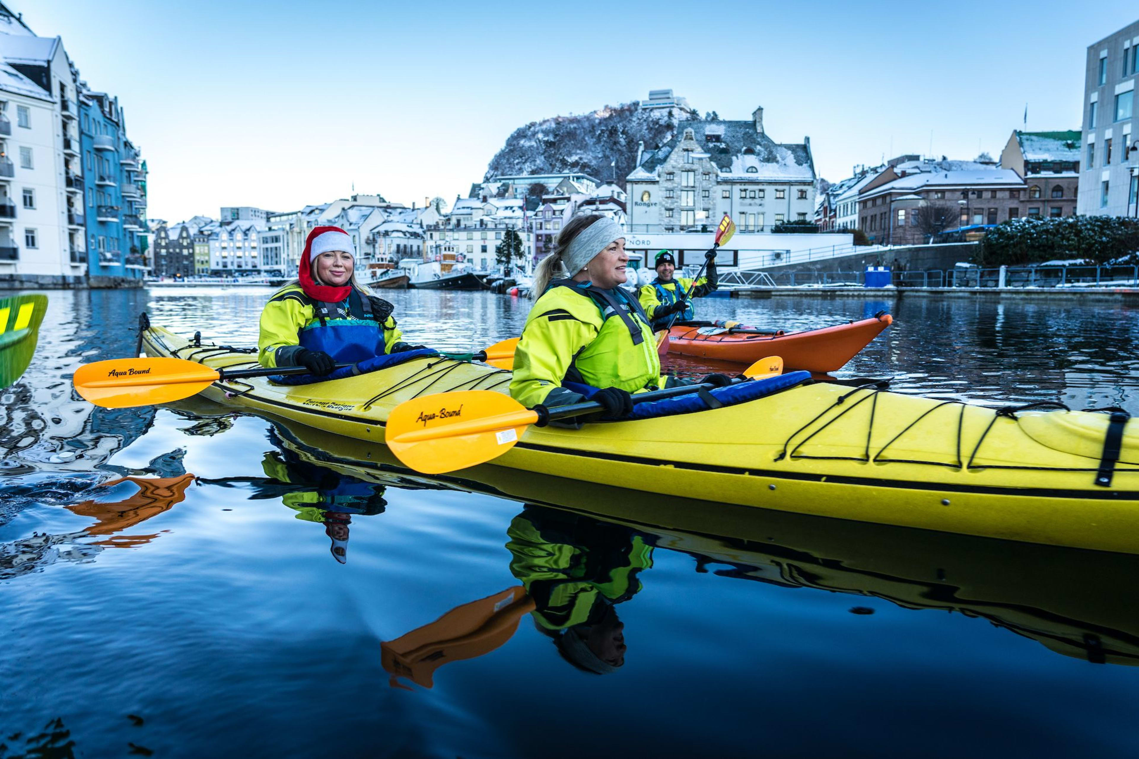Three people kayaking in the water, surrounded by snow-covered buildings, with bright yellow and blue life jackets.