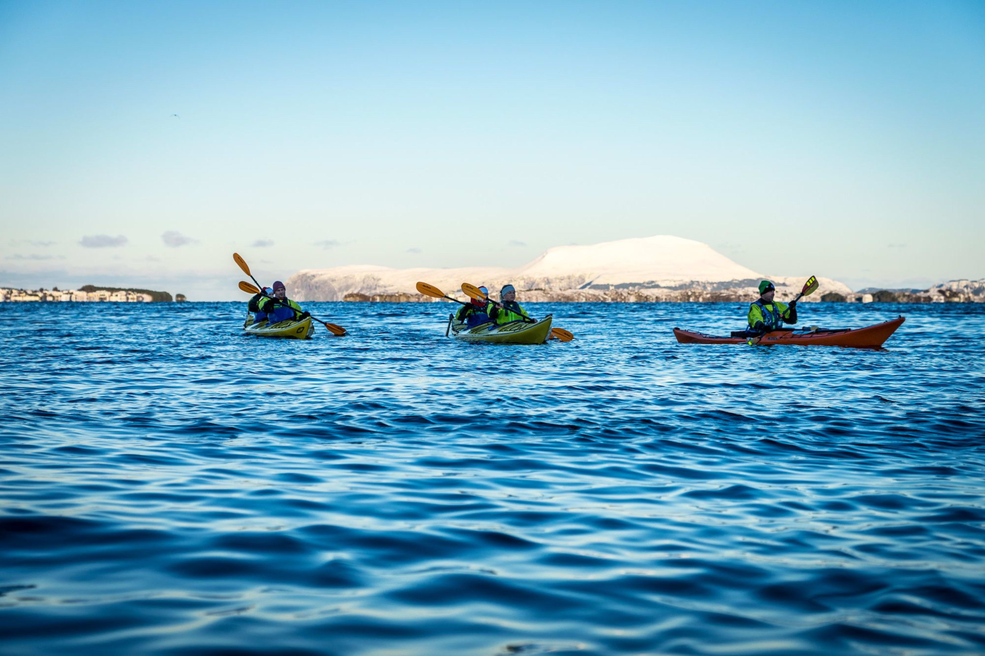 Five people kayaking on calm blue water, surrounded by snow-capped mountains and buildings along the shoreline.